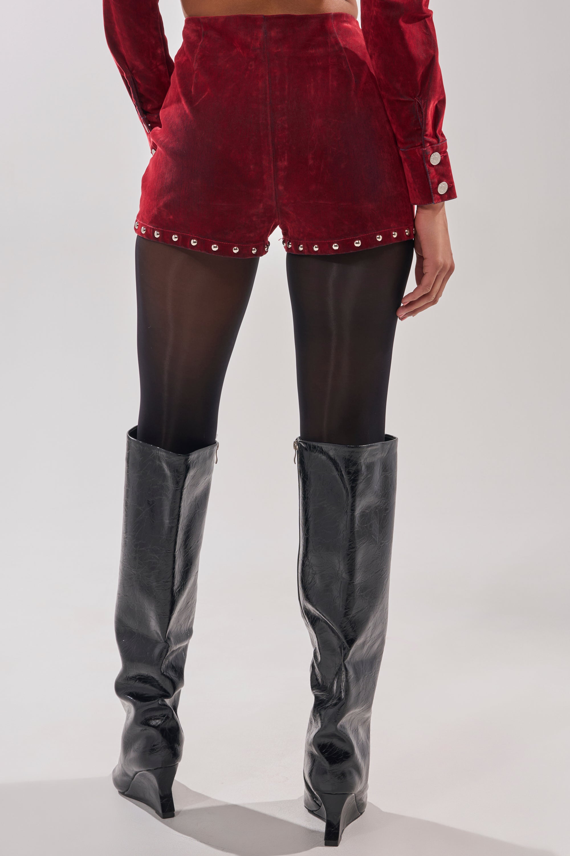 A person wears black knee-high boots, sheer black tights, and the RODEO VIBES VELVET DENIM STUDDED SHORT in red velvet with silver studs along the hem. Their hands rest at their sides against a plain light gray background.