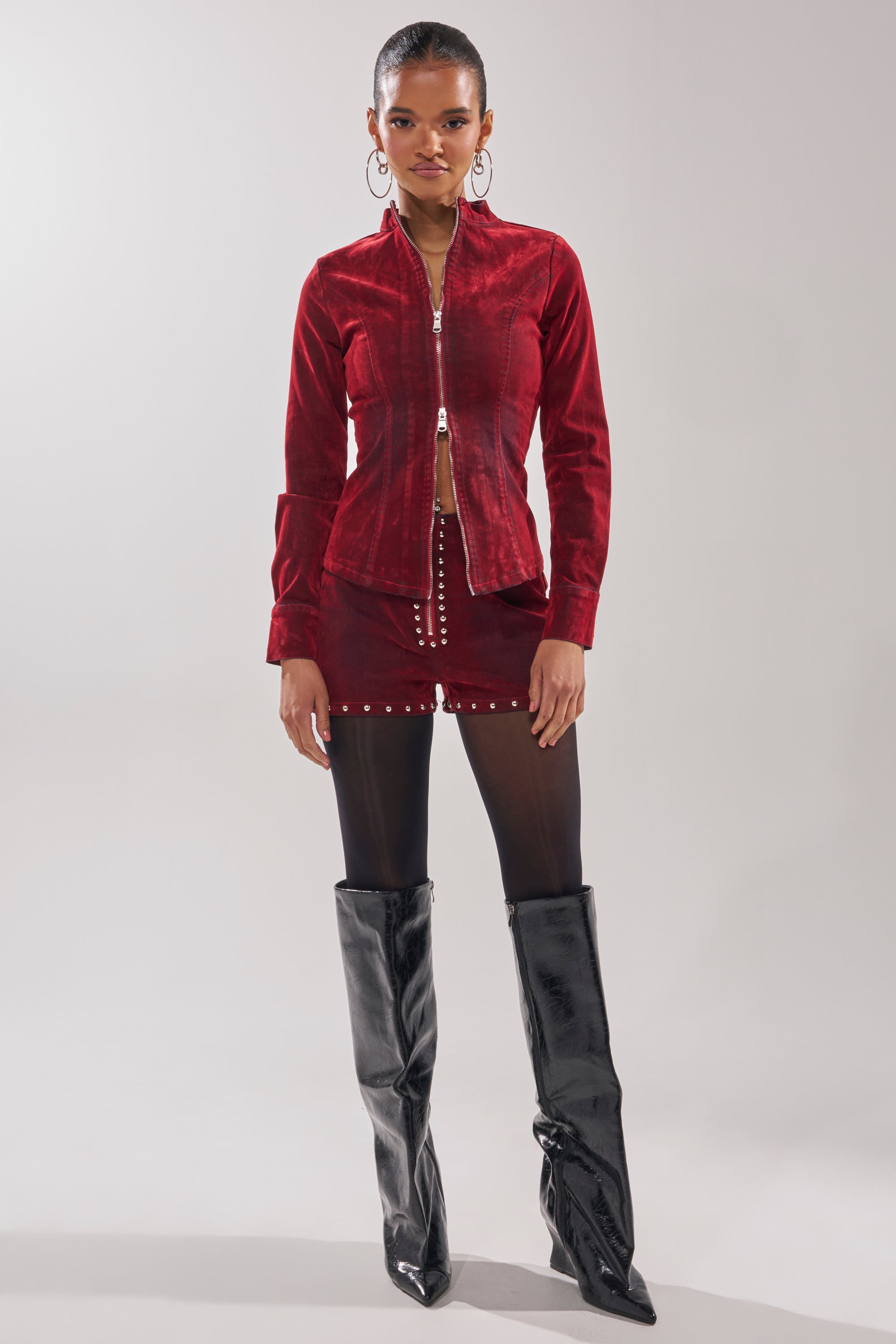 A woman poses against a plain background wearing the RODEO VIBES VELVET DENIM STUDDED SHORT with a matching red velvet zip-up jacket, black tights, large hoop earrings, and tall black patent leather boots.