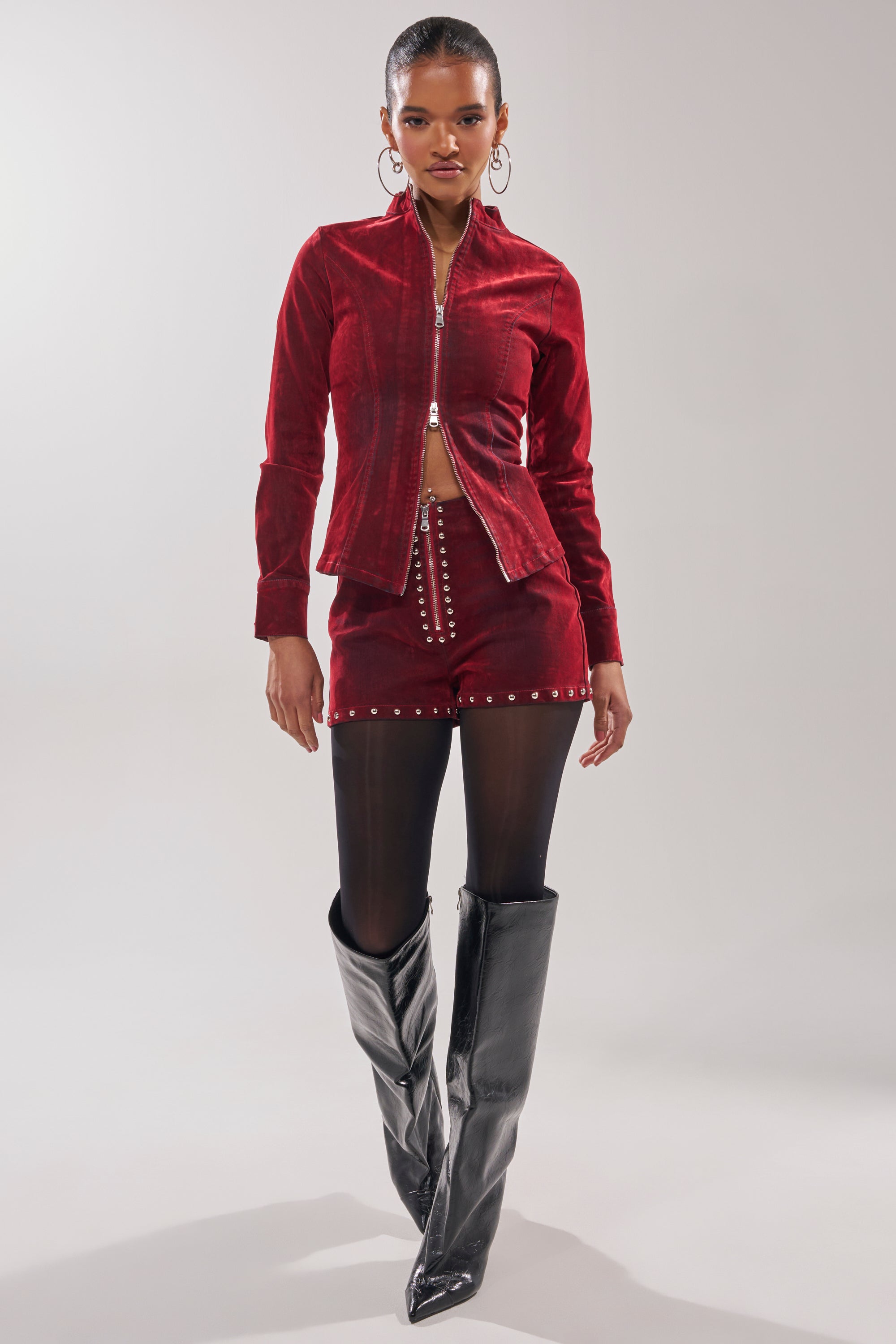 A woman confidently poses against a plain light background in the RODEO VIBES VELVET DENIM STUDDED SHORT with a matching red velvet zip-up jacket, black tights, and shiny black knee-high boots.