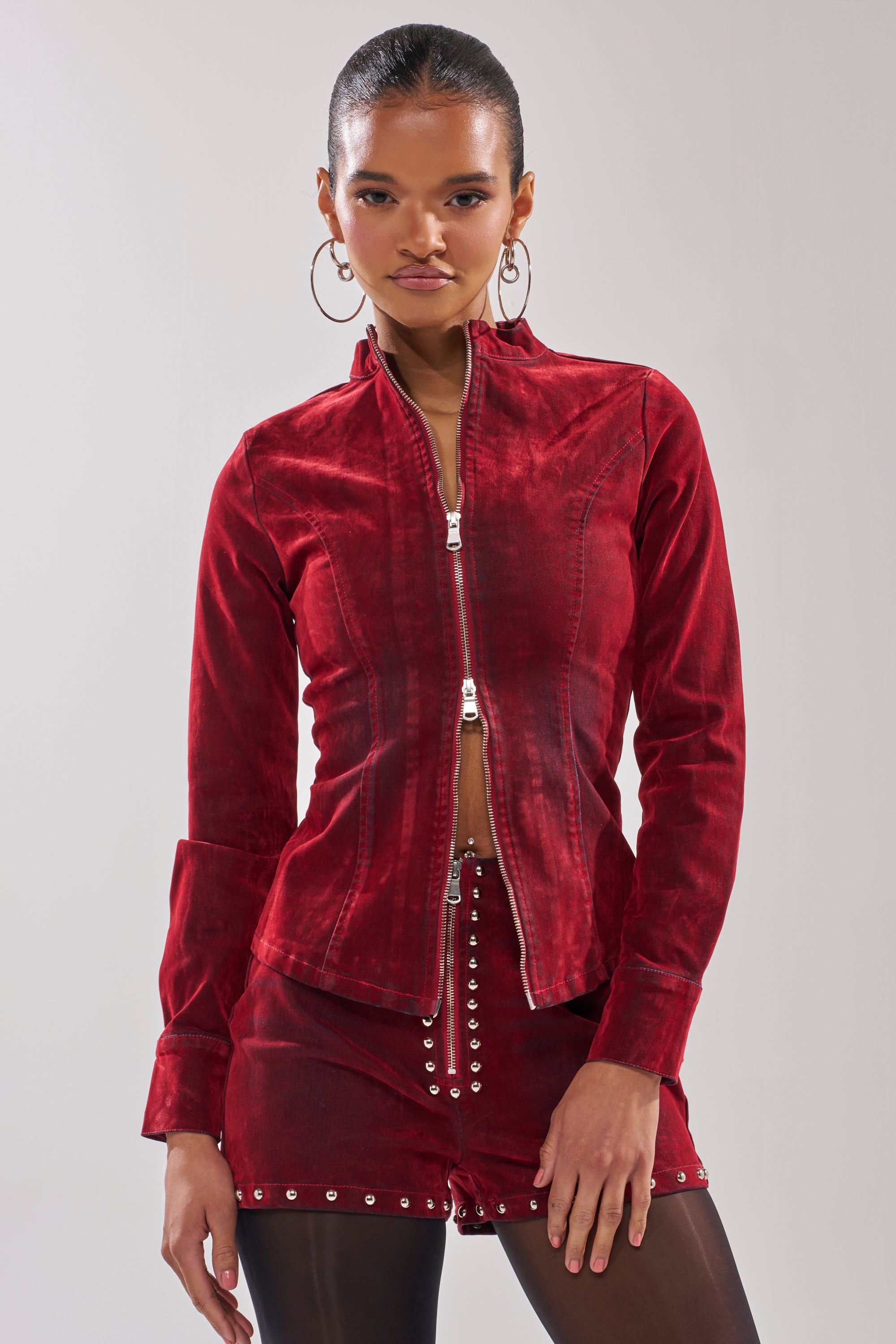 A woman sports the RODEO VIBES VELVET DENIM ZIP UP jacket with matching red studded shorts, accessorized with hoop earrings and dark tights, channeling a modern cowgirl look against a plain light background.