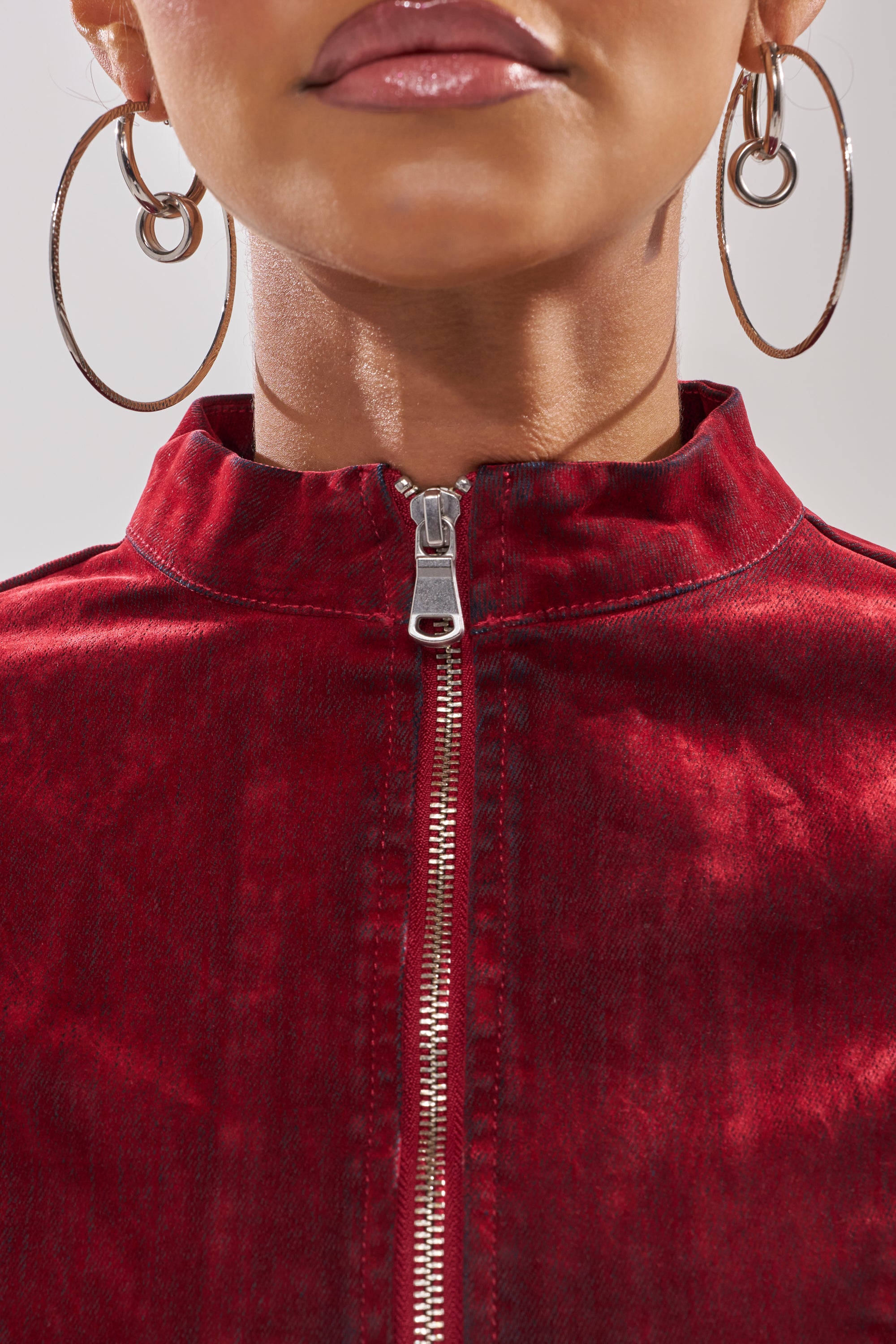 A close-up shows a person in large silver hoop earrings wearing the RODEO VIBES VELVET DENIM ZIP UP, a red velvet jacket with a high collar. Only the lower face and upper chest appear in the image.
