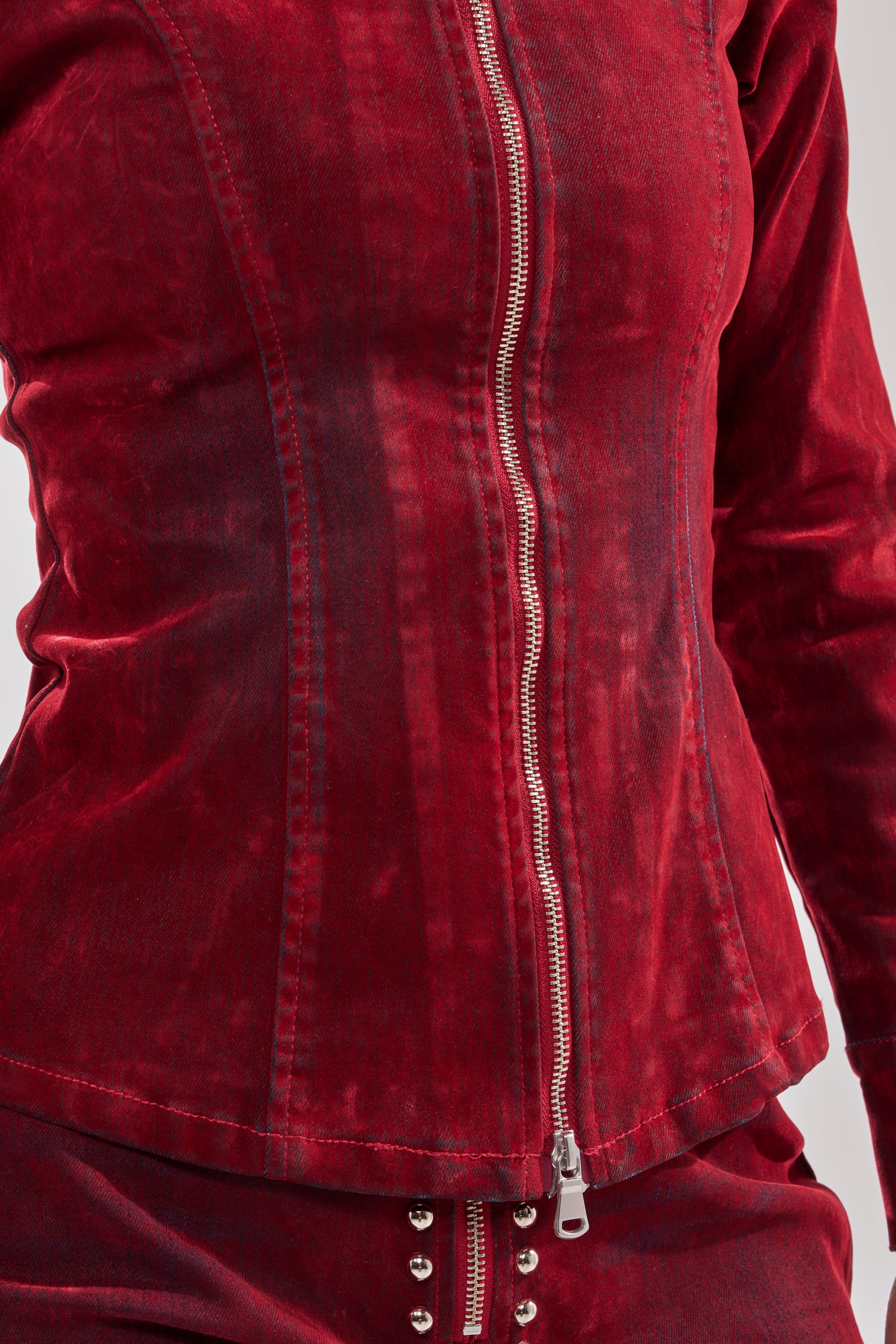 A close-up of a person wearing the RODEO VIBES VELVET DENIM ZIP UP in red, featuring a silver zipper and matching velvet pants with silver studded waistband, set against a plain light background.