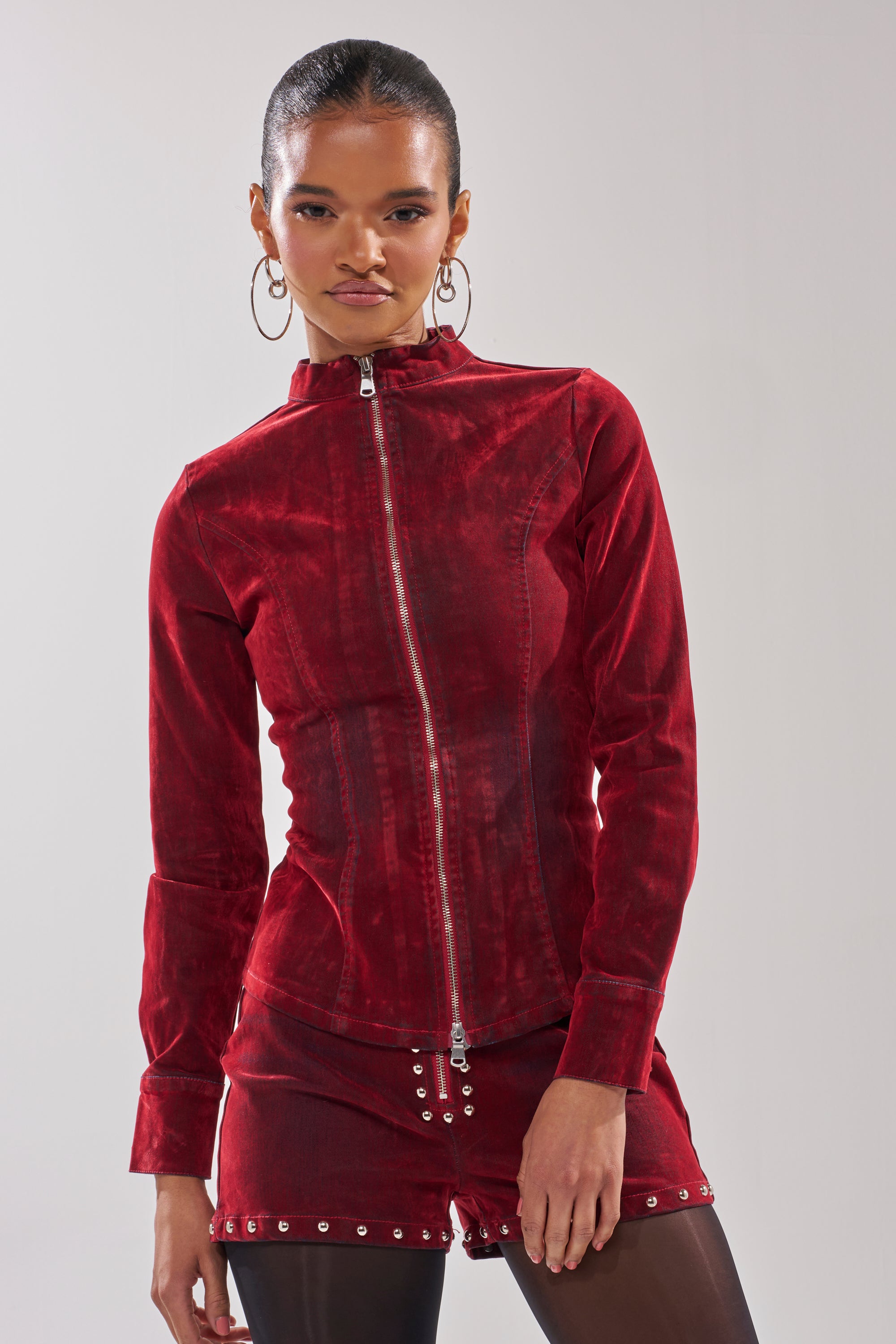 A woman with hoop earrings wears the RODEO VIBES VELVET DENIM ZIP UP, a tailored fit, long-sleeve red velvet zip-up jacket paired with matching red shorts with silver accents, standing against a plain light background.