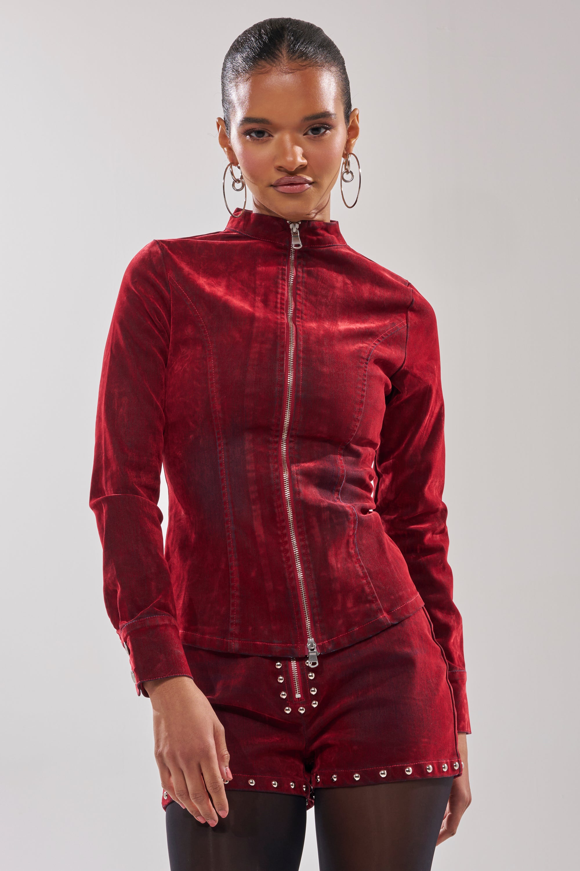 A woman faces forward in the RODEO VIBES VELVET DENIM ZIP UP, a tailored red velvet zip-up jacket with matching studded shorts and large hoop earrings, set against a plain light gray background.