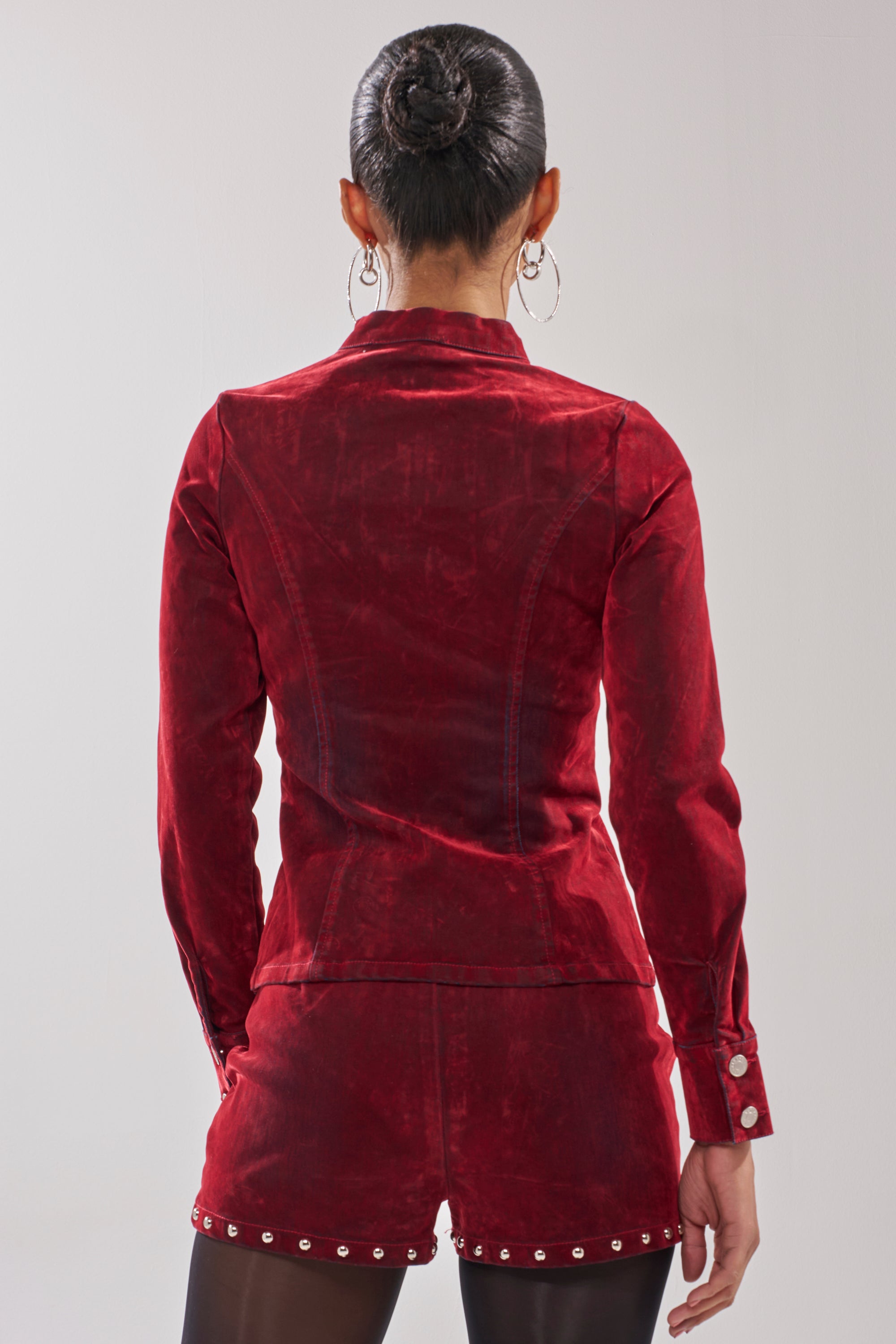 Seen from the back against a plain background, a person with their hair in a bun and large hoop earrings wears the RODEO VIBES VELVET DENIM ZIP UP jacket and matching shorts, both in red velvet with silver-studded hems.