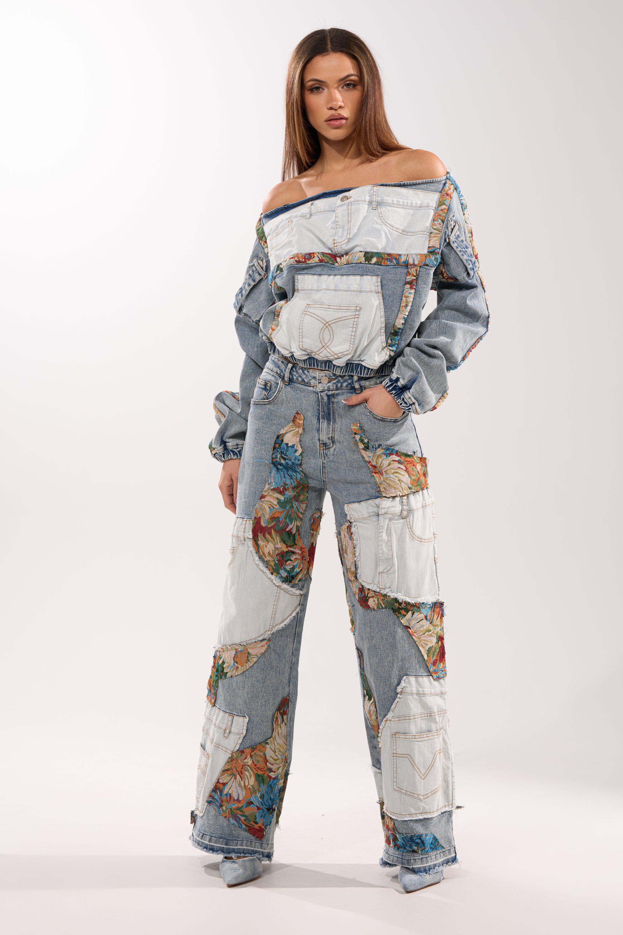 A woman confidently poses against a plain light background wearing the CHANGE IT UP WIDE LEG DENIM—a matching mixed-wash set with white patchwork, floral details, off-the-shoulder long sleeves, and high-rise wide-leg jeans.