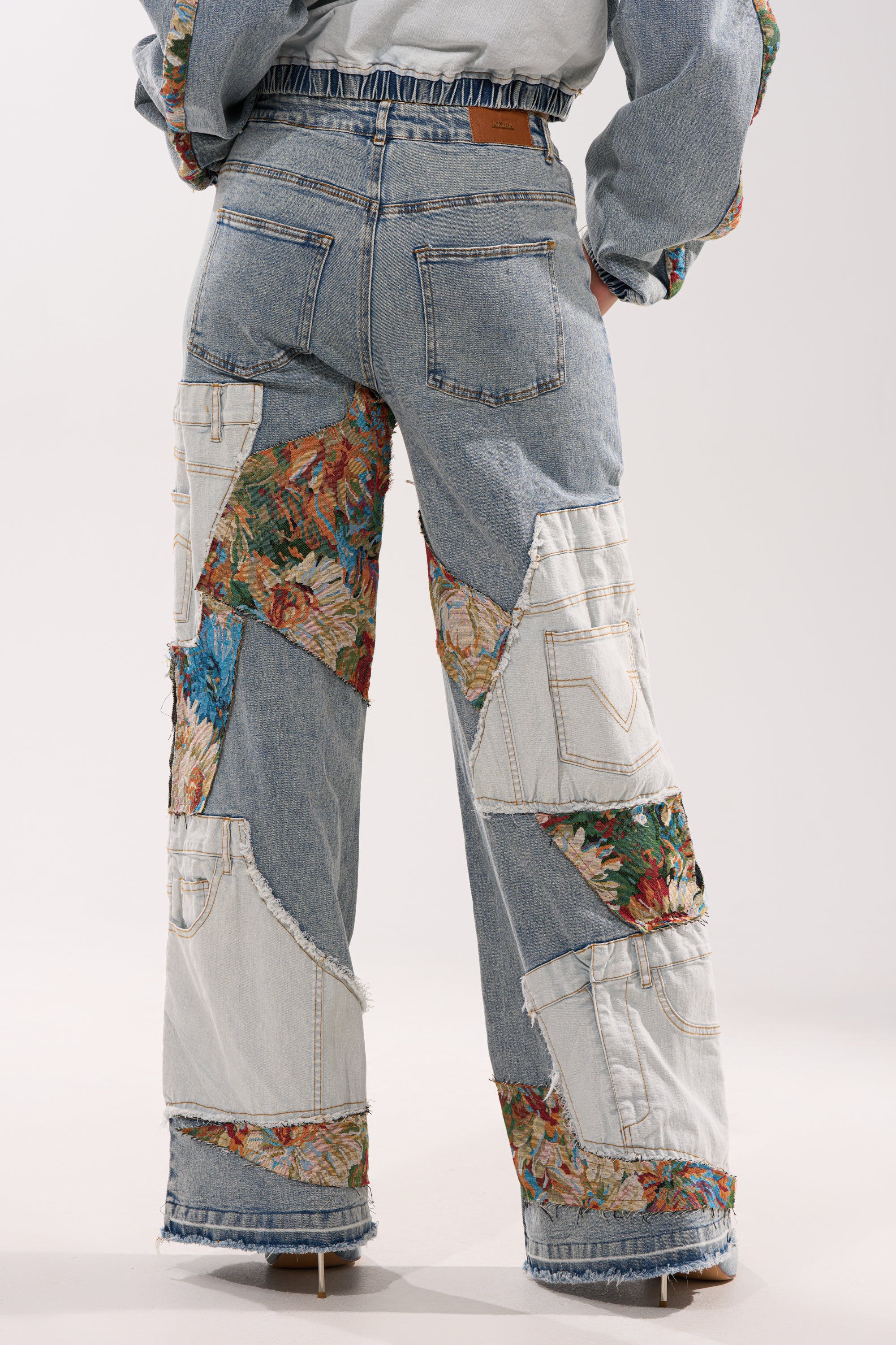 Someone wears the CHANGE IT UP WIDE LEG DENIM, showcasing patchwork mixed-wash denim and floral fabric, standing with their back to the camera against a plain background for a cool, vintage-inspired look.