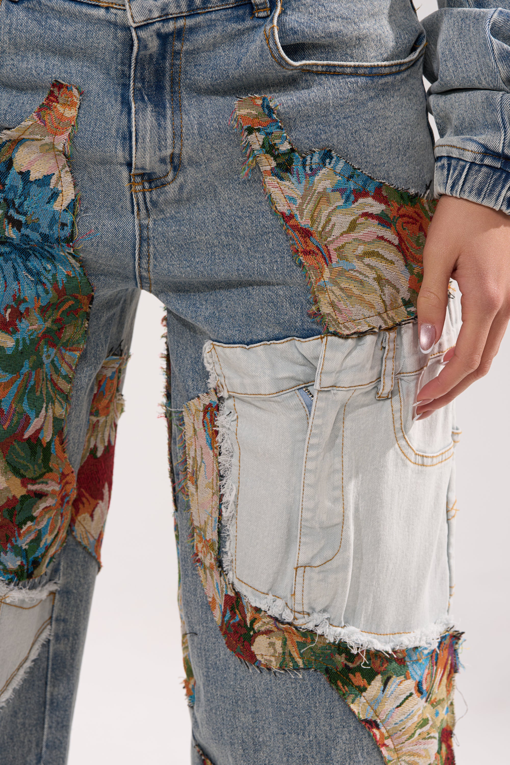 A close-up of a person wearing CHANGE IT UP WIDE LEG DENIM, featuring large colorful floral patches and frayed edges. A manicured hand rests near the pocket on these mixed-wash patchwork jeans.