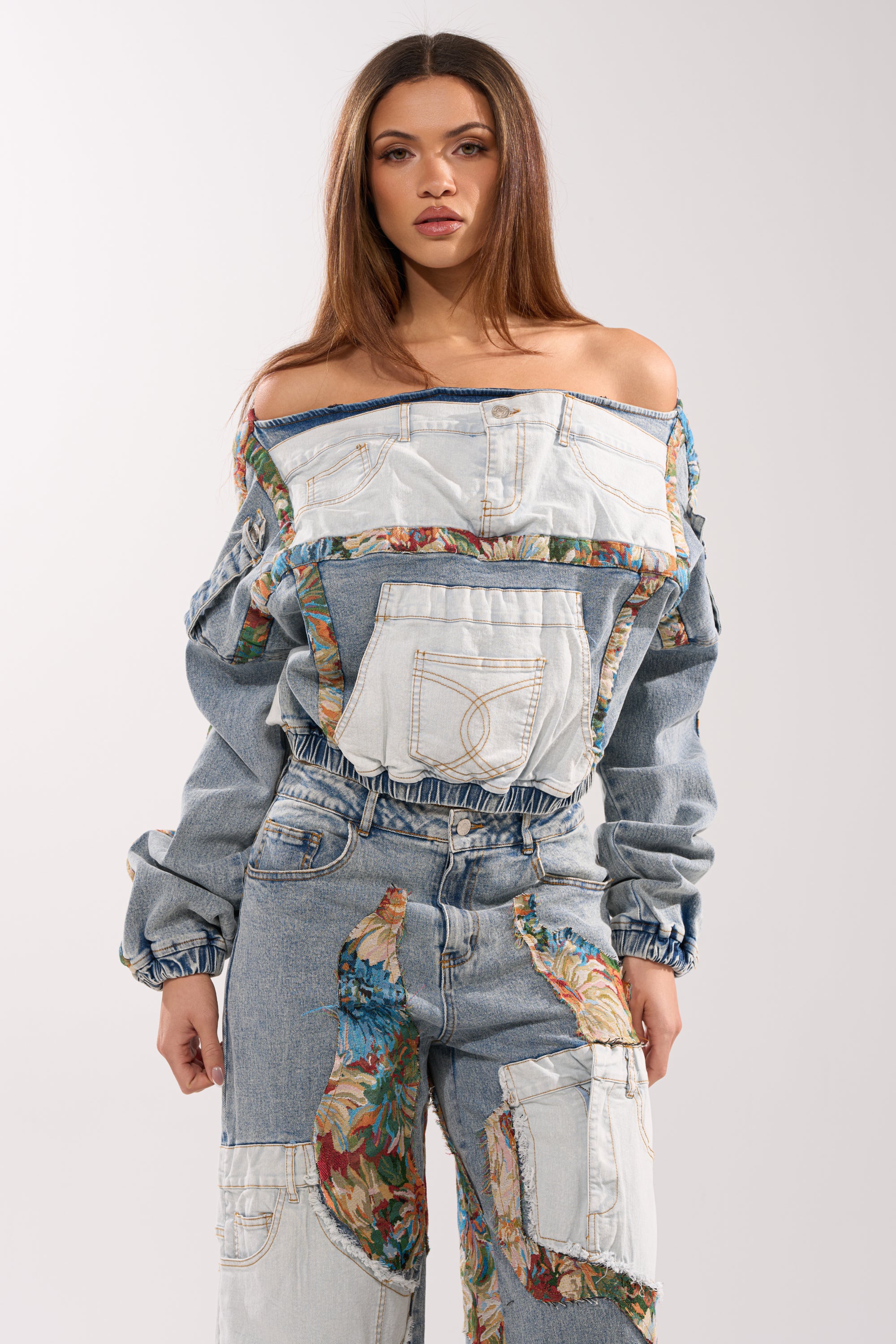 A woman with long brown hair models the CHANGE IT UP DENIM SWEATSHIRT, featuring patchwork denim, exposed seams, floral fabric accents, and multiple visible pockets against a plain light background.
