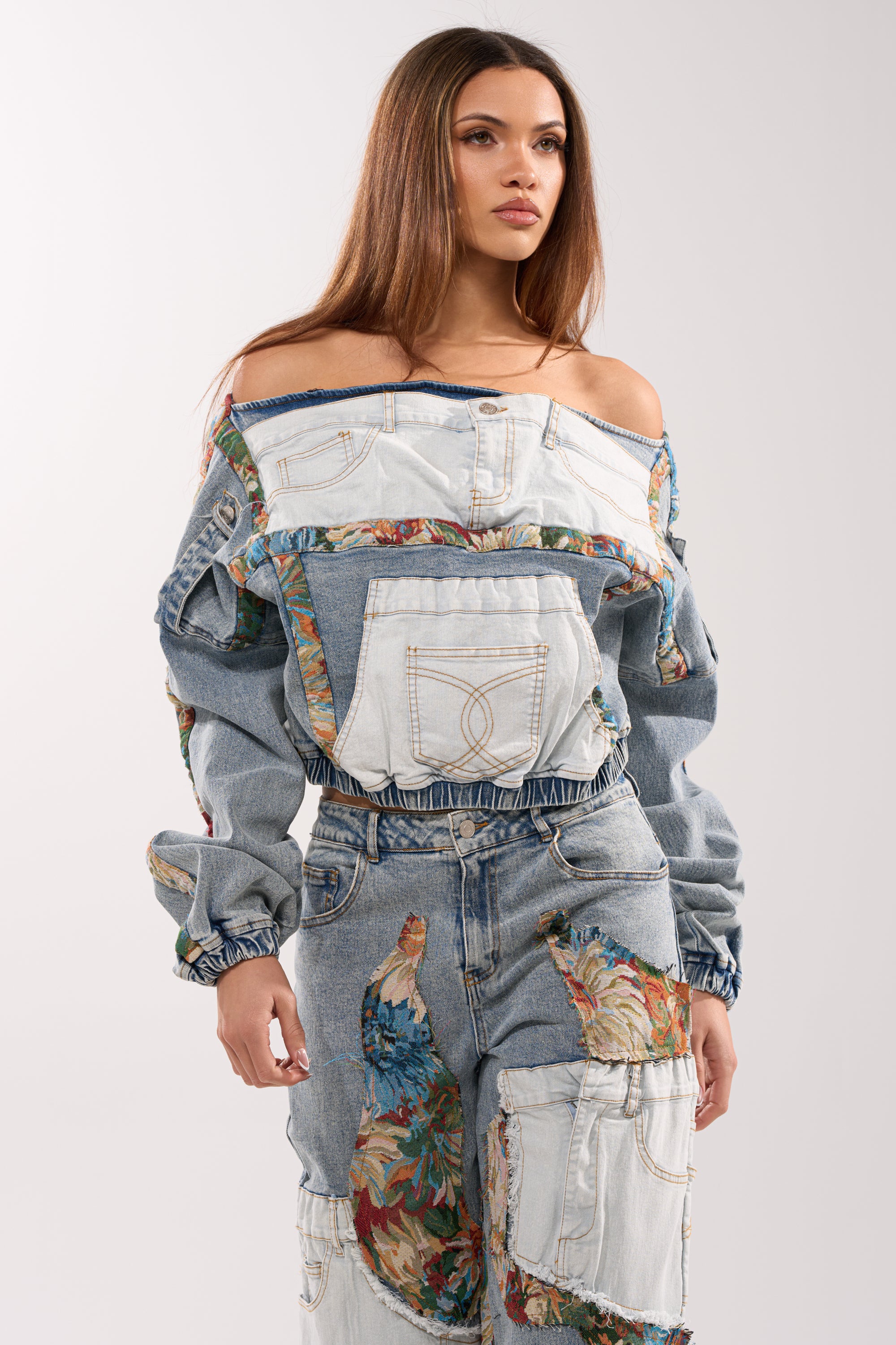 A woman models the CHANGE IT UP DENIM SWEATSHIRT, a street-style off-the-shoulder top with patchwork designs, floral accents, and visible pockets, paired with matching denim pants against a plain light background.