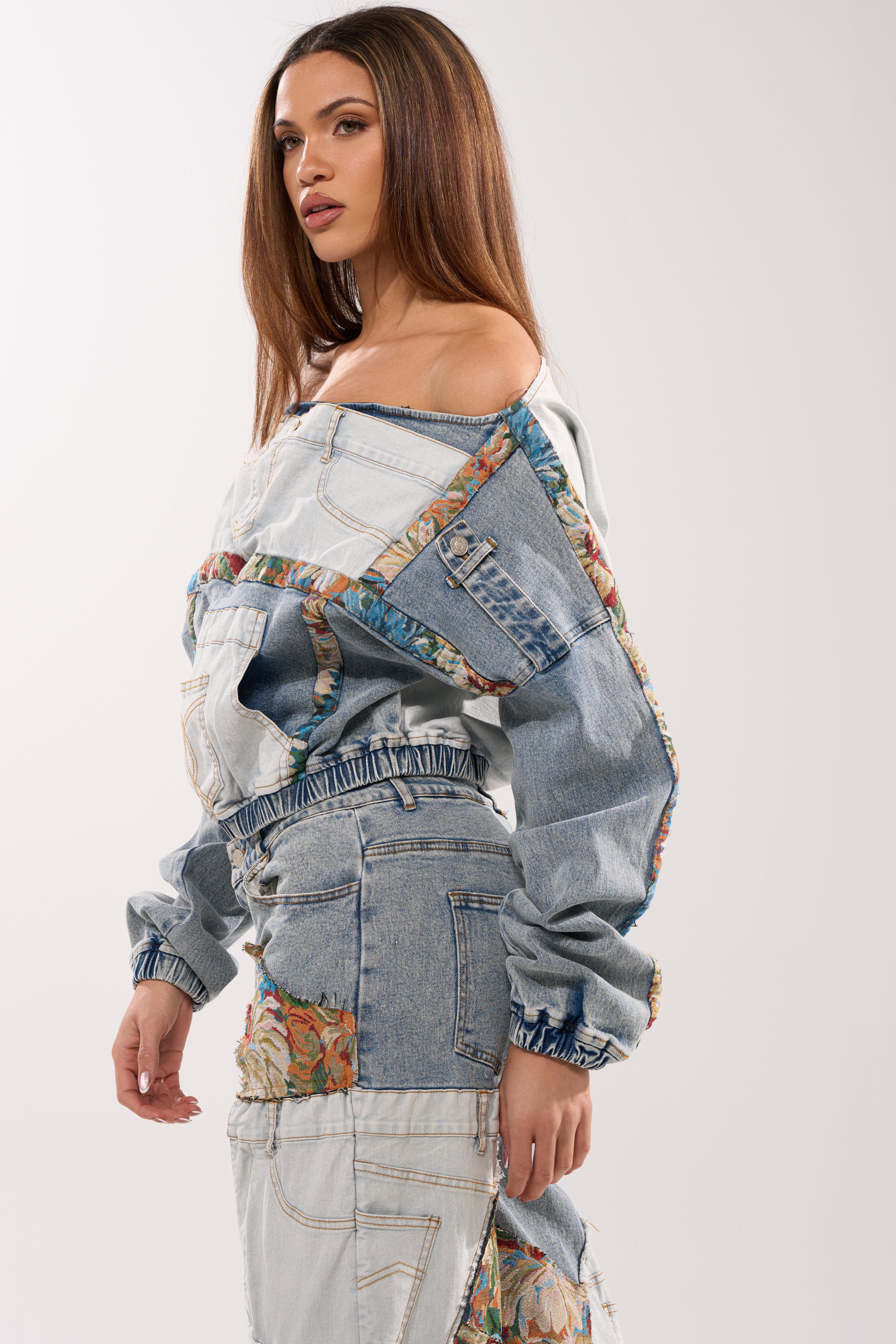 A woman with long brown hair wears the CHANGE IT UP DENIM SWEATSHIRT with floral fabric accents, styled streetwear fashion, and poses against a plain light background.