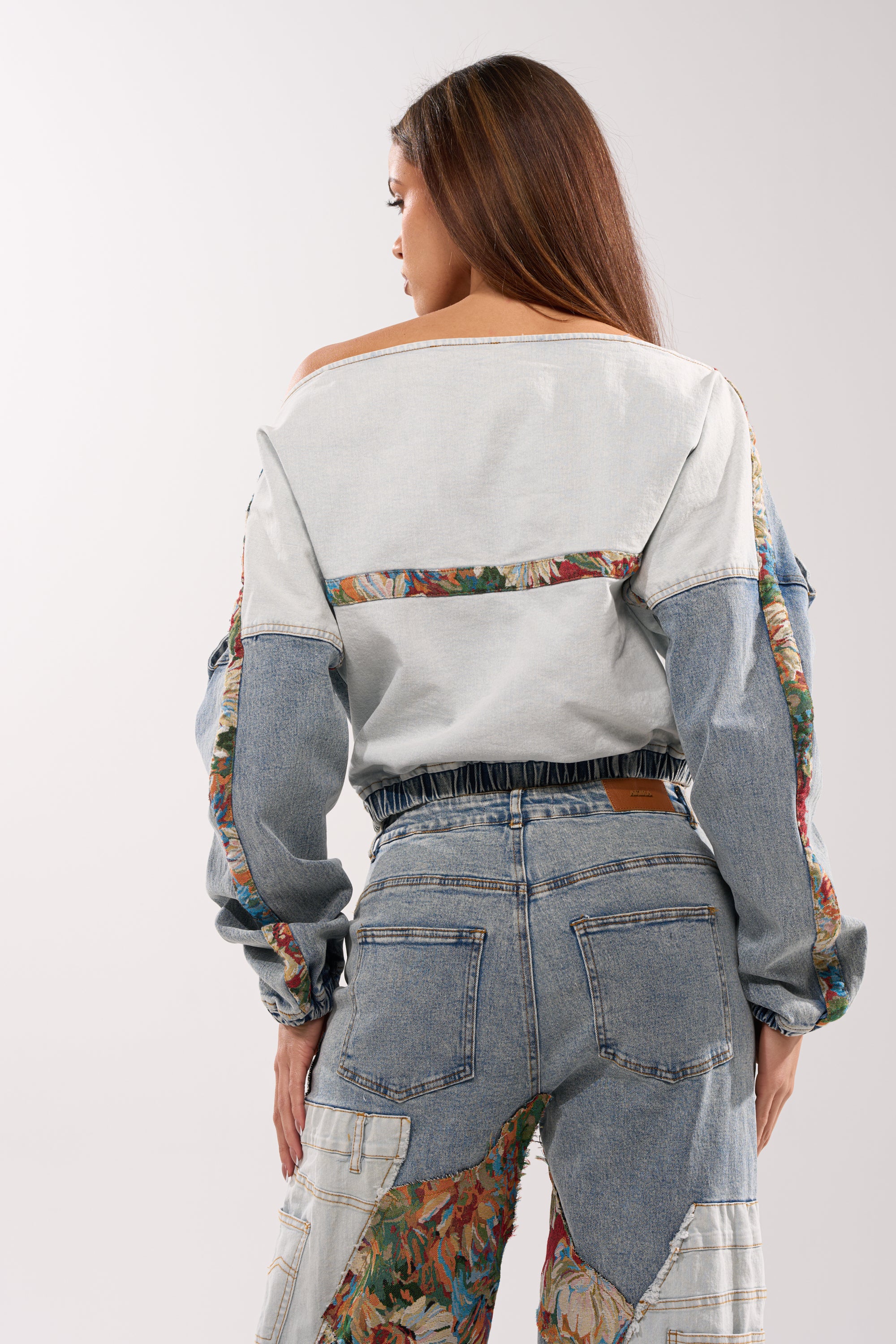 A woman with long brown hair stands facing away, wearing the CHANGE IT UP DENIM SWEATSHIRT and matching jeans with colorful floral accents, styled in streetwear fashion against a plain background.
