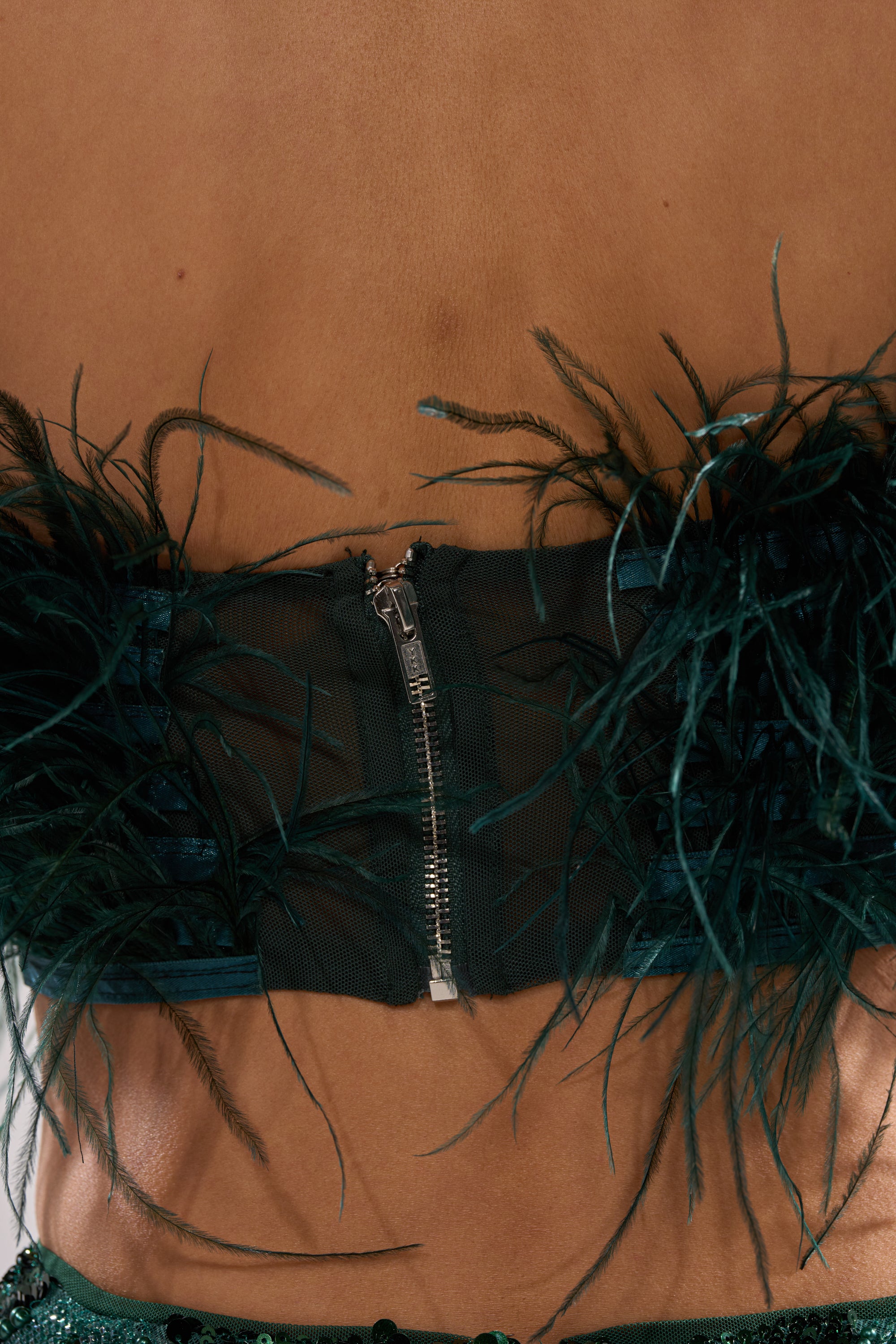Close-up of a person's bare back wearing the FLY AWAY FEATHER BRA TOP—a dark green crop top featuring sheer black mesh, a visible center zipper, and decorative feathers outlining the edges for a striking night-out look.