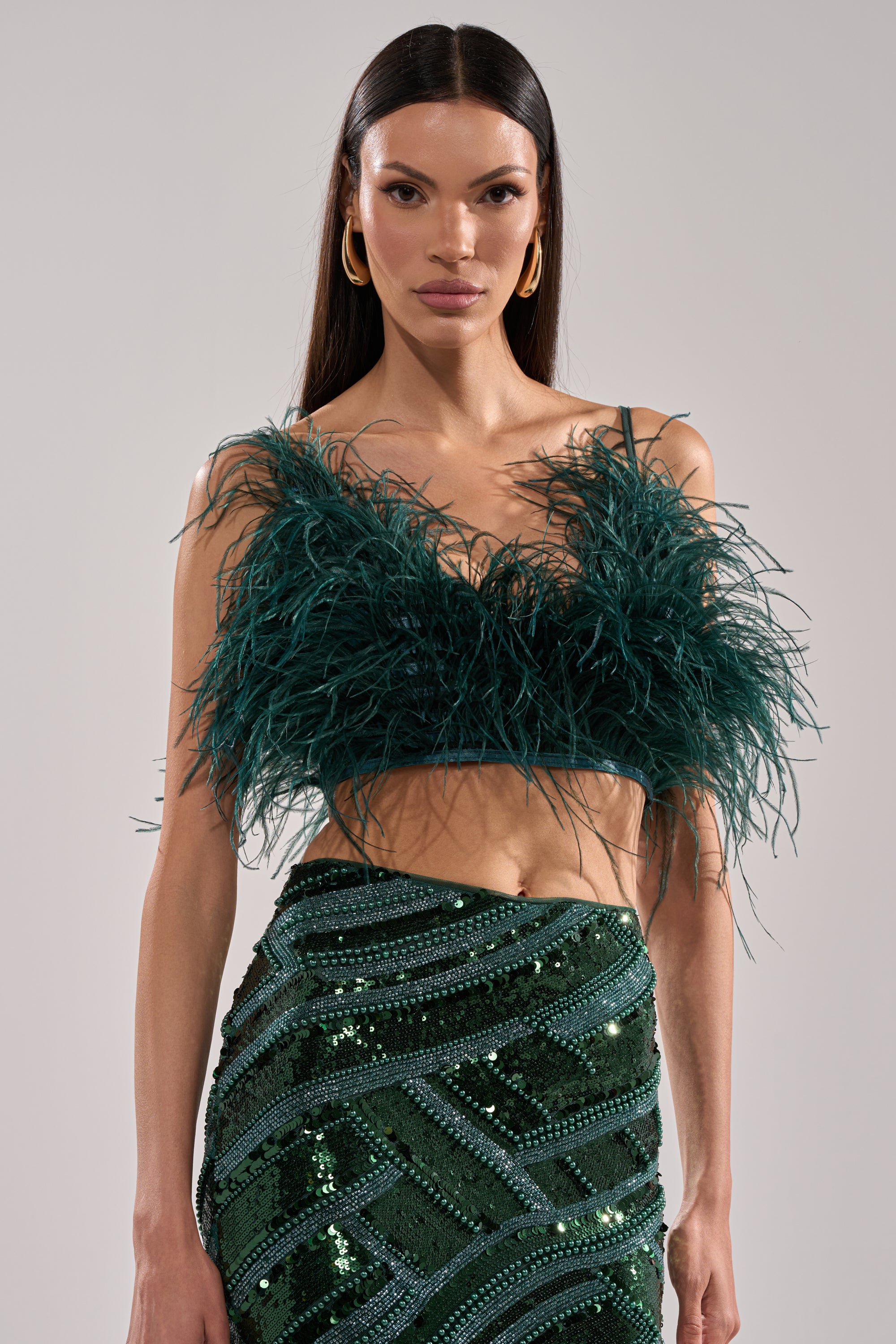 A woman with long straight hair models the FLY AWAY FEATHER BRA TOP in dark green, paired with a green sequin skirt featuring diagonal stripes, as she poses confidently against a plain backdrop.