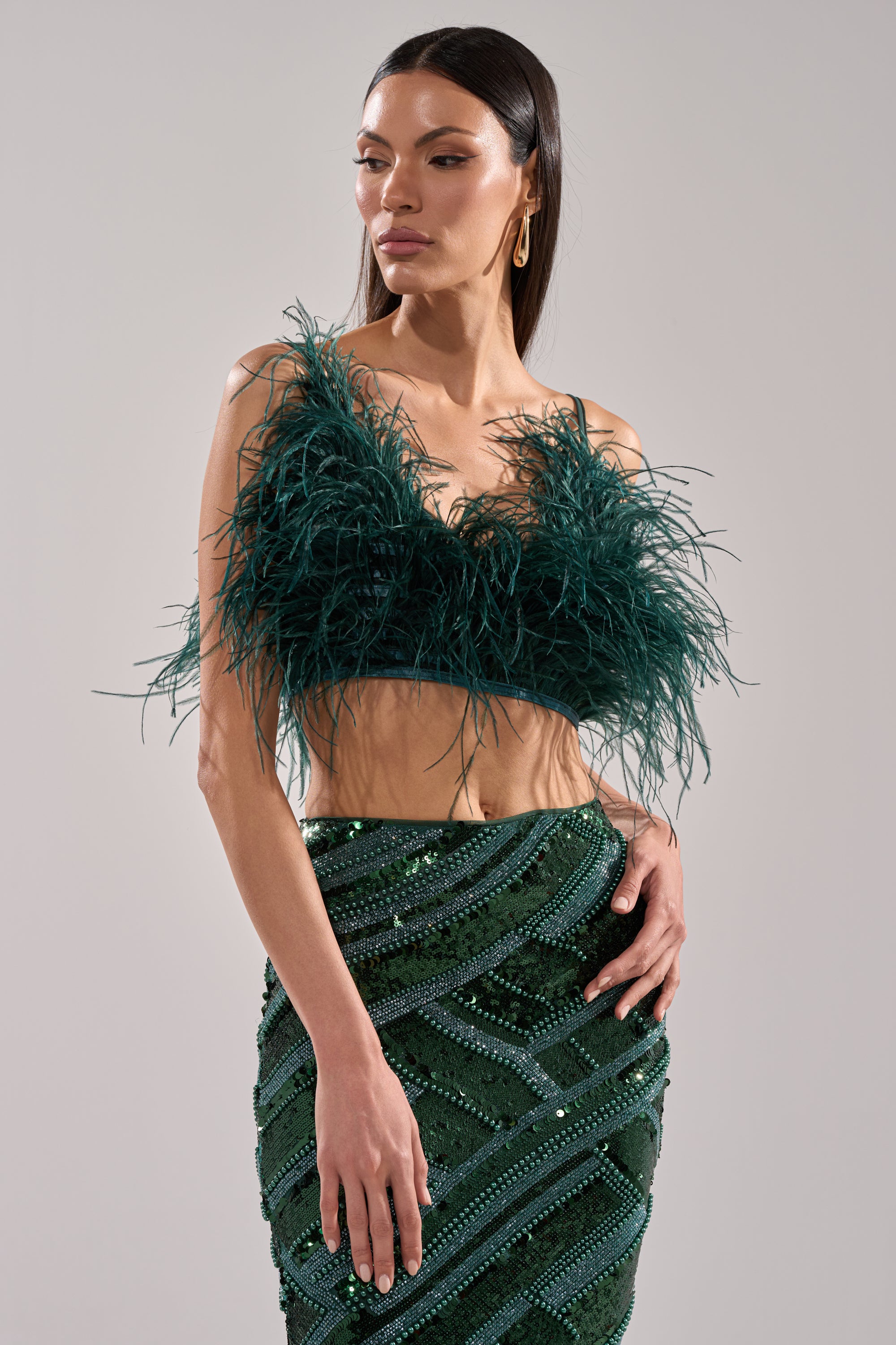 A woman models the FLY AWAY FEATHER BRA TOP, paired with a beaded, sequined green skirt, standing confidently against a light gray background—a bold and stylish statement look.