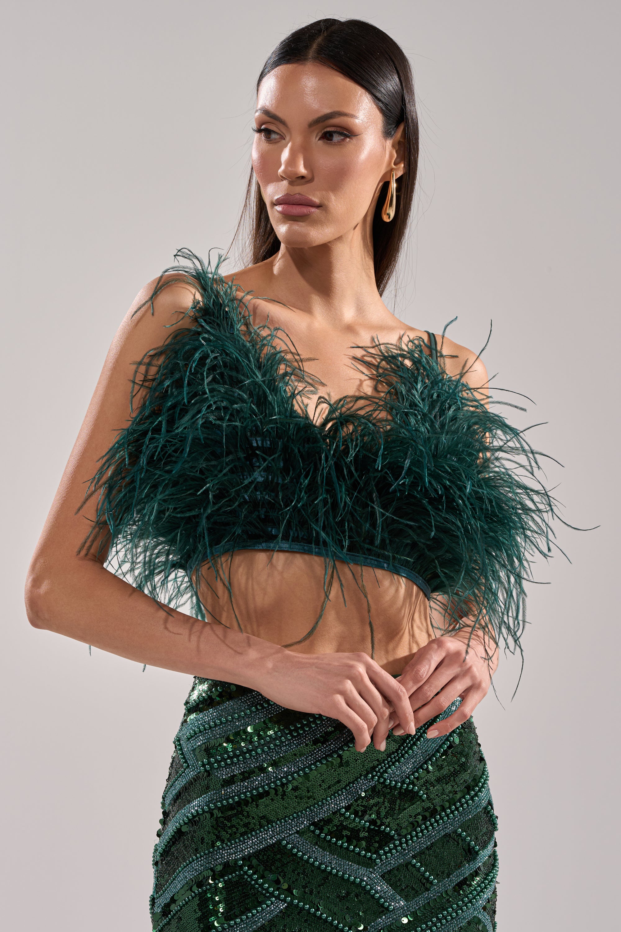 Wearing the FLY AWAY FEATHER BRA TOP in dark green and a green sequined skirt, a woman poses with one hand on her hip, gazing to the side against a plain light gray background—a true statement piece.