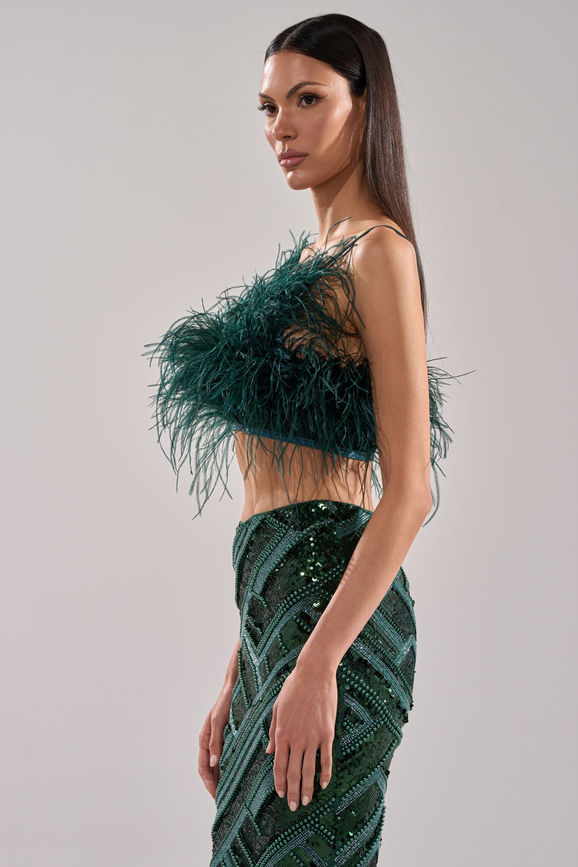 A woman in the FLY AWAY FEATHER BRA TOP and a high-waisted, sequined, geometric-patterned green skirt stands in profile against a plain light gray background—this eye-catching look is a true statement piece.
