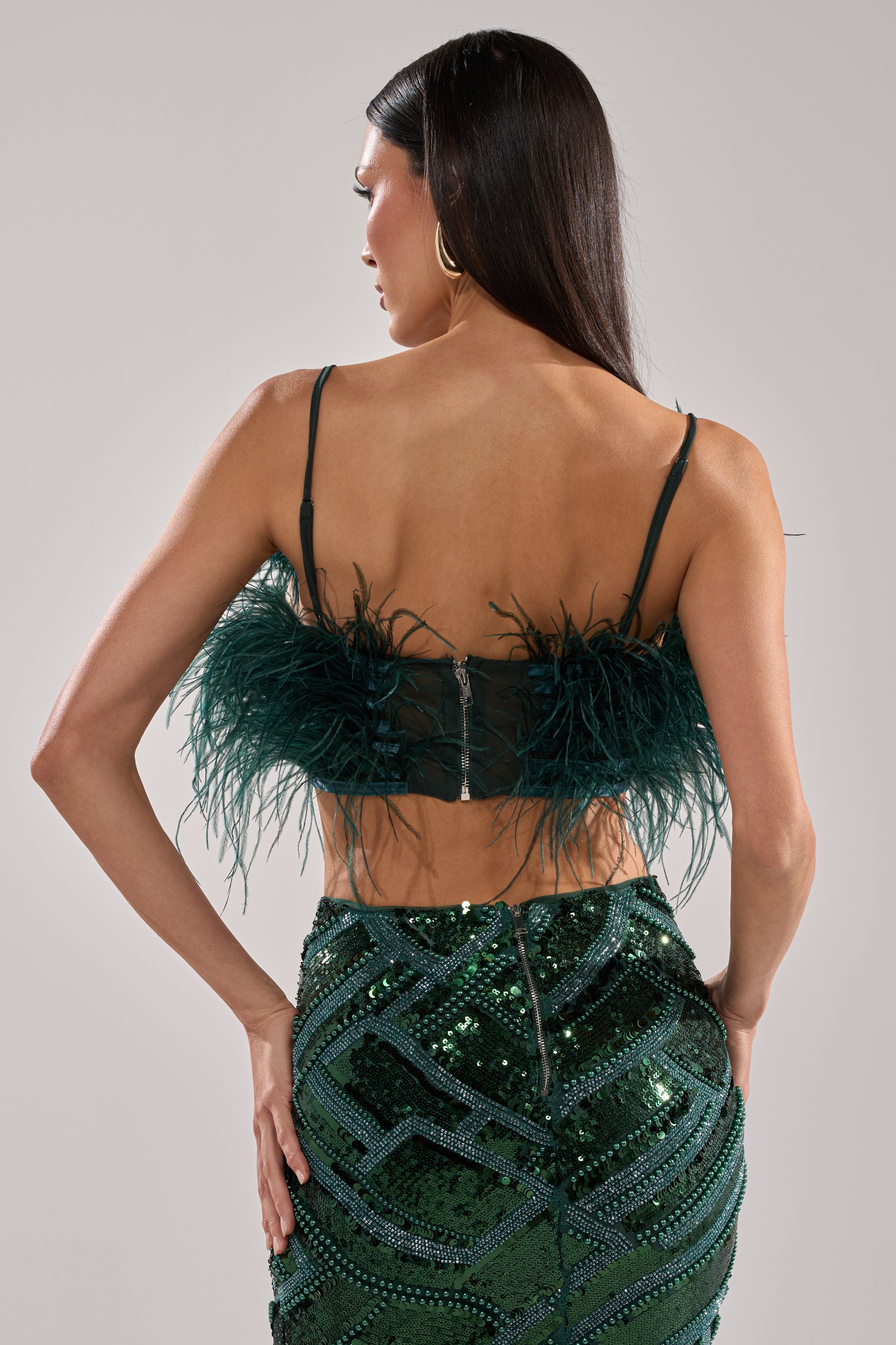 A woman stands with her back to the camera, modeling the FLY AWAY FEATHER BRA TOP in green paired with a shiny green sequined skirt against a plain light gray background—a bold night-out statement look.