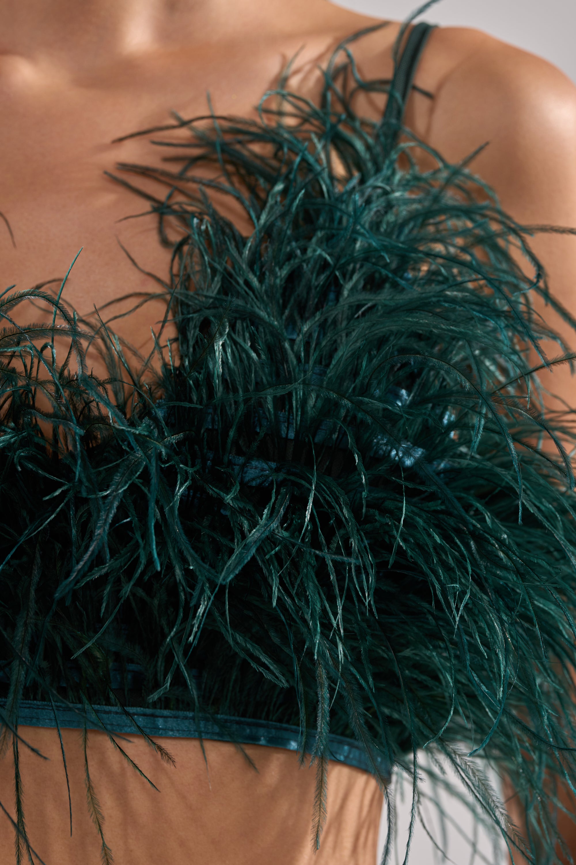 A close-up of someone wearing the FLY AWAY FEATHER BRA TOP in dark green with thin straps, featuring detailed textured feathers across the chest for an eye-catching look.