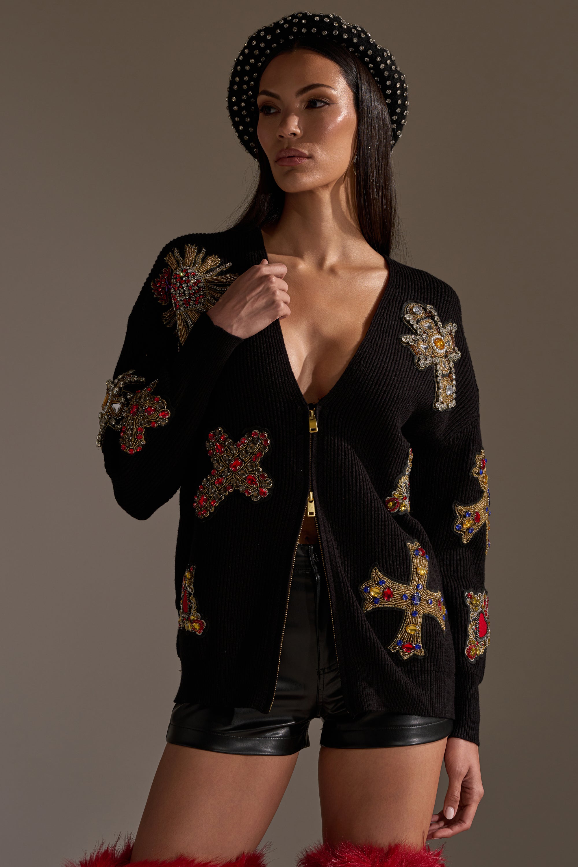 A woman wearing the FEELING EXTRA EMBELLISHED SWEATER with jeweled cross designs, paired with black leather shorts, a beaded black beret, and furry red boots, stands confidently against a plain background. 0
