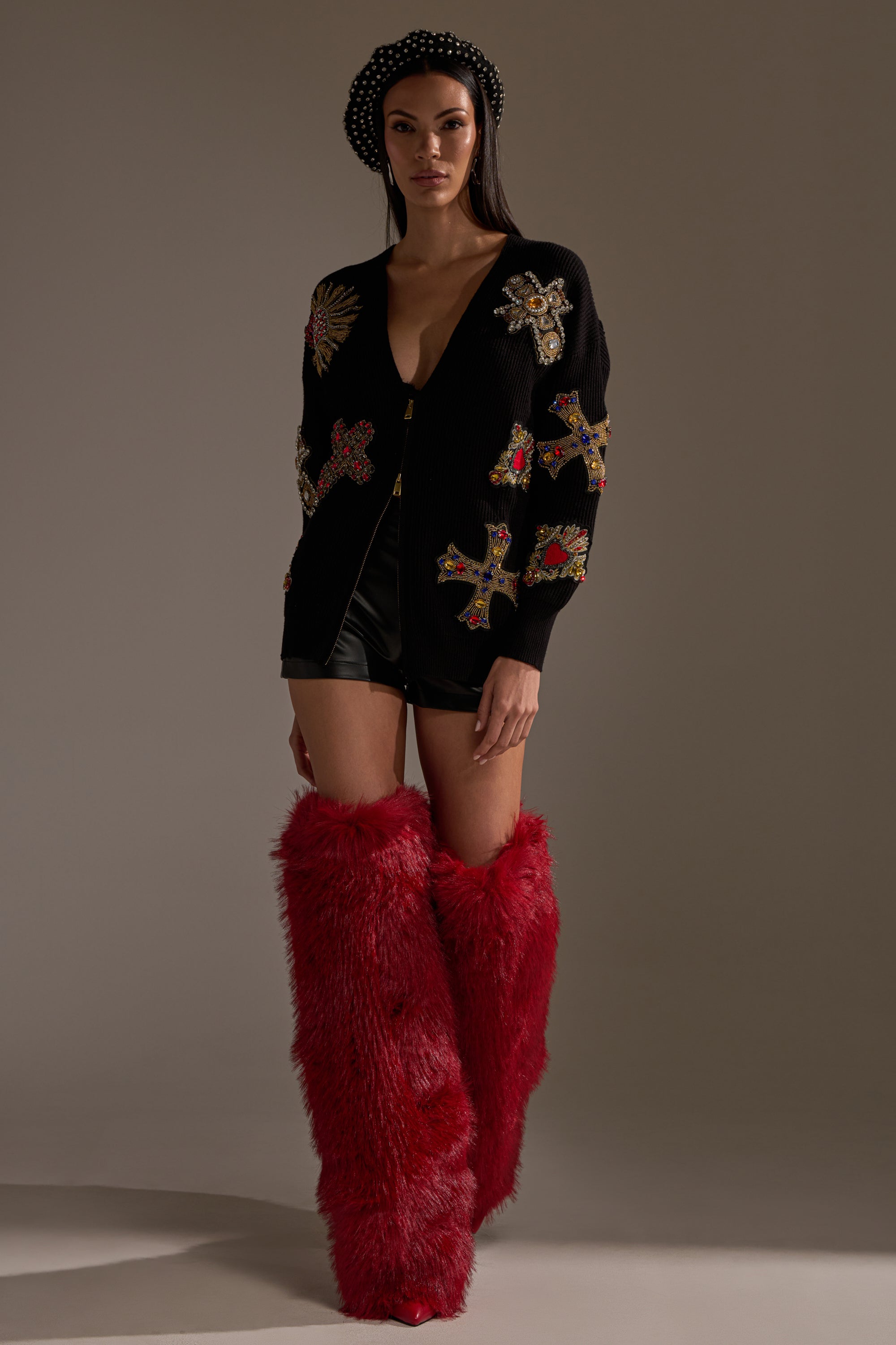 A woman wears the FEELING EXTRA EMBELLISHED SWEATER with beaded cross designs, paired with black shorts, a black polka dot beret, and tall red furry boots, standing against a plain neutral background. 9