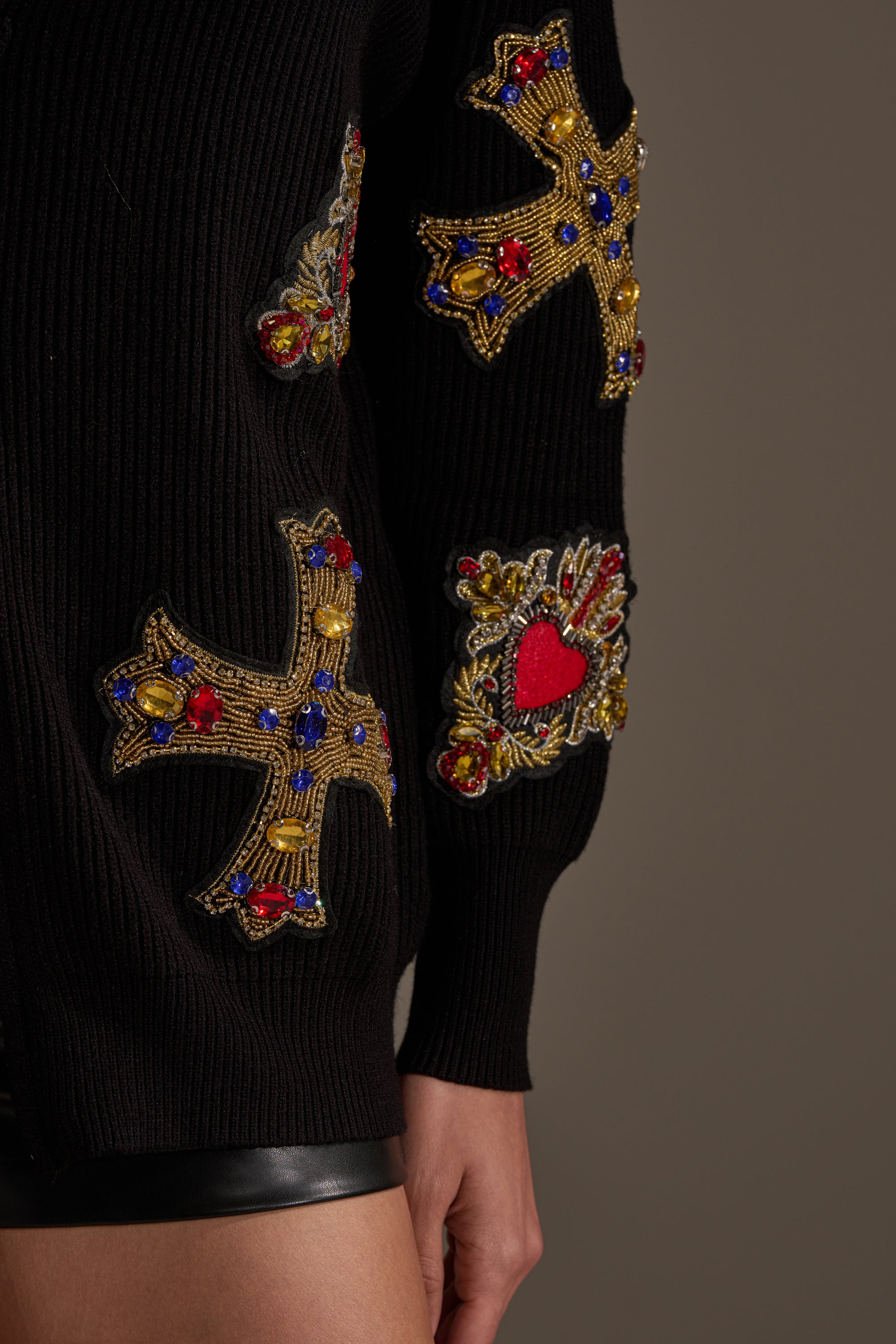 A close-up of someone wearing the FEELING EXTRA EMBELLISHED SWEATER, featuring ornate embroidered gold crosses, a red heart with red, blue, and gold beading, and ribbed knit texture on the arm and upper leg. 11