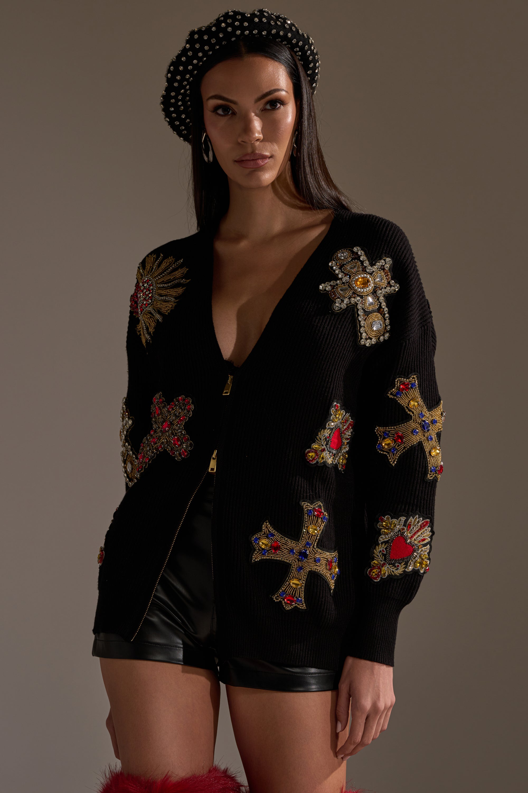 A woman poses against a softly lit background wearing the FEELING EXTRA EMBELLISHED SWEATER with jeweled crosses, a black beret with pearls, black leather shorts, and red feathered boots. 1