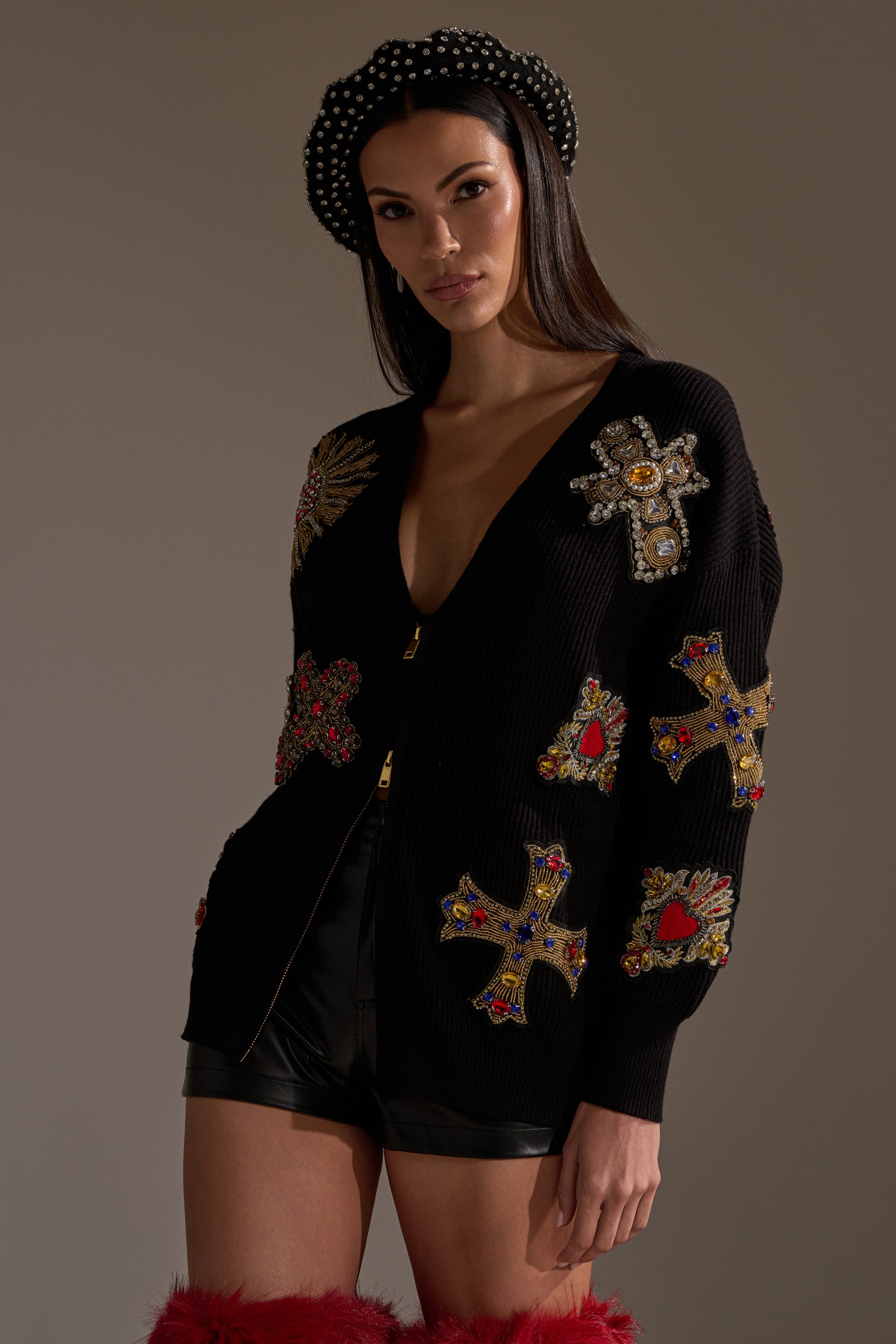 A woman wears the FEELING EXTRA EMBELLISHED SWEATER with jeweled cross patterns, black leather shorts, a black polka dot beret, and red furry boots, posing against a plain background. 2