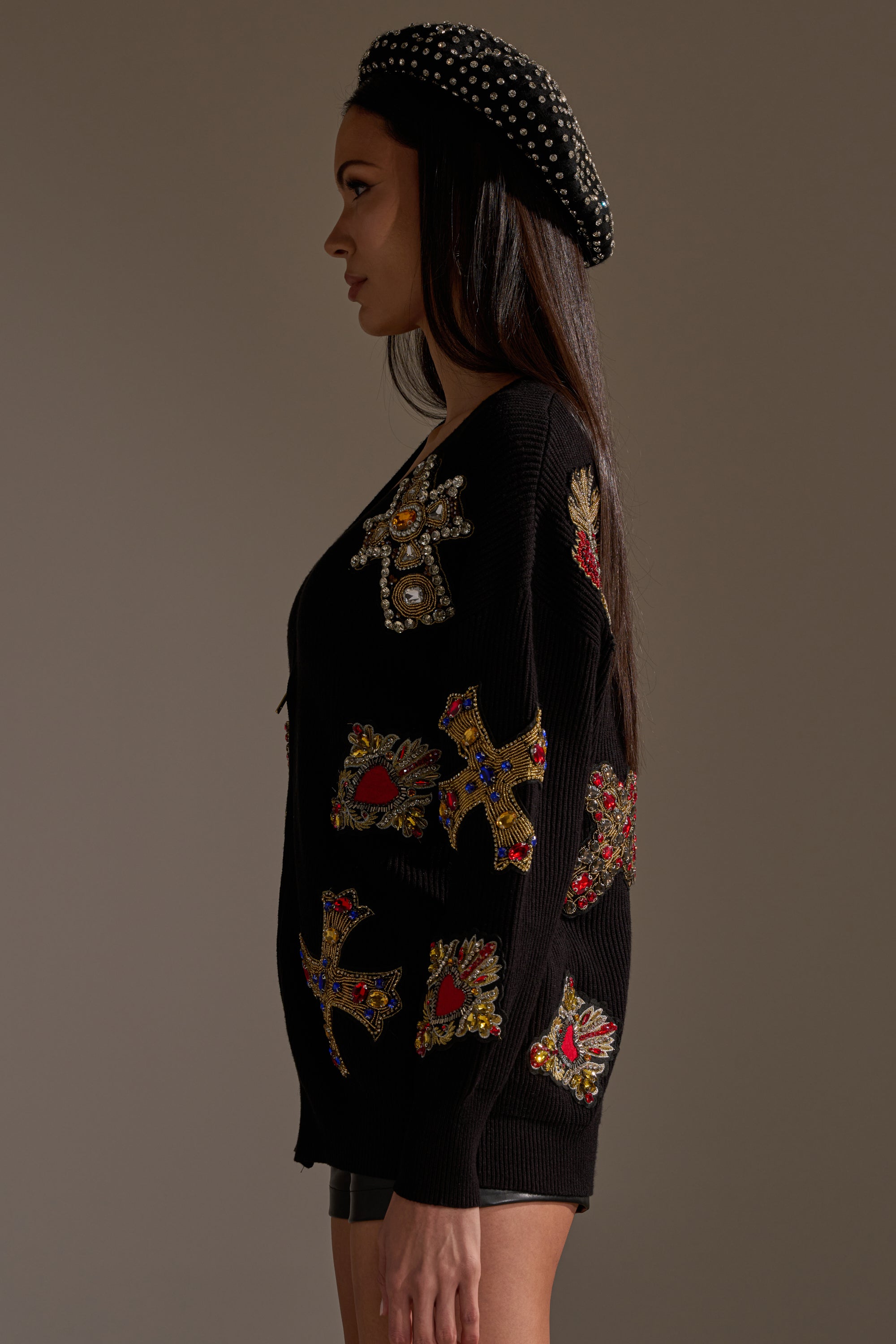 A woman in profile wears the FEELING EXTRA EMBELLISHED SWEATER with colorful jeweled cross and heart patches, paired with black shorts and a beret featuring silver studs. The ribbed knit adds texture against the neutral background. 3