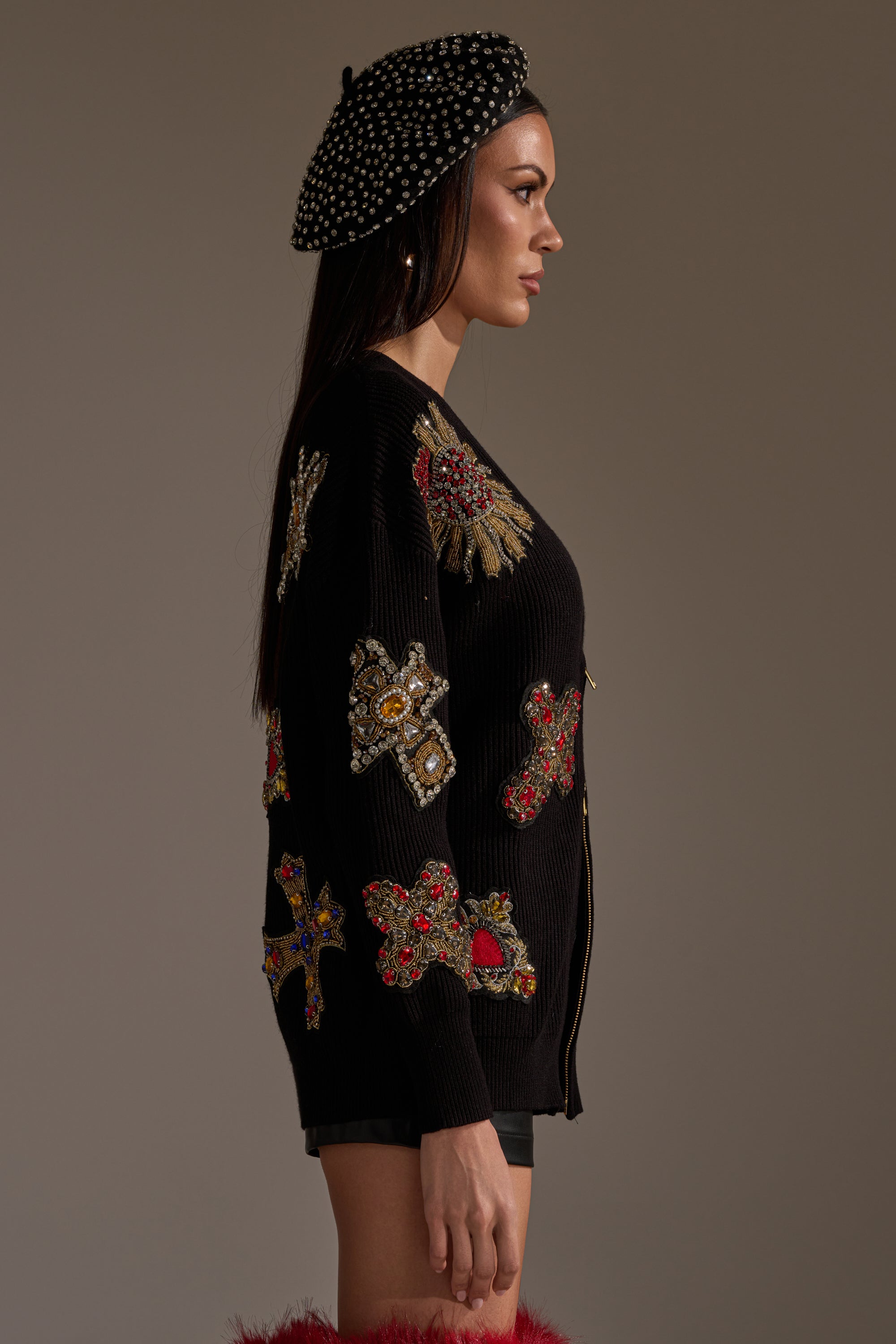 A woman in profile wears the FEELING EXTRA EMBELLISHED SWEATER with jeweled cross designs, a pearl-detailed black beret, hoop earrings, and black shorts against a plain, neutral background. 4