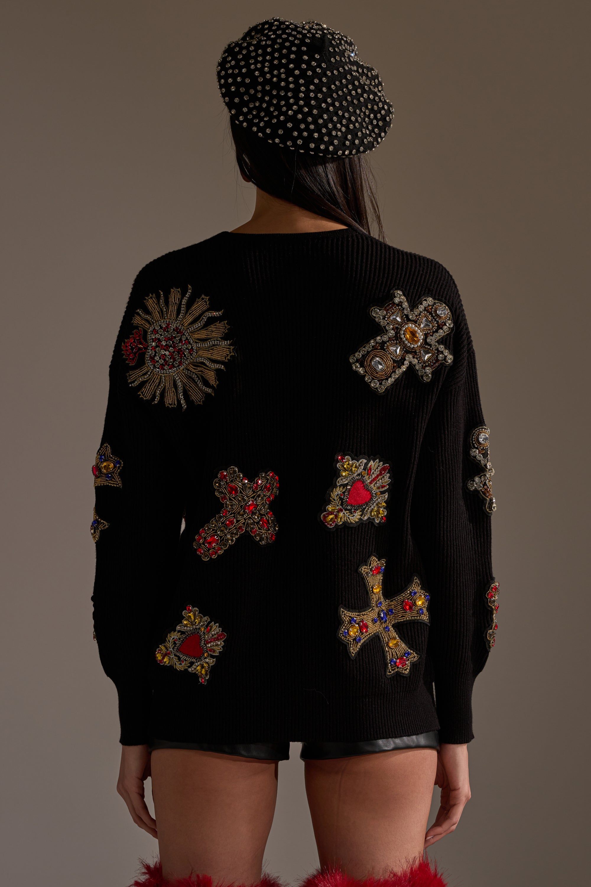 A person faces away, wearing the FEELING EXTRA EMBELLISHED SWEATER—a black ribbed knit with colorful, ornate cross-shaped embroidered patches—paired with black leather shorts, a studded black beret, and red furry boots. 5