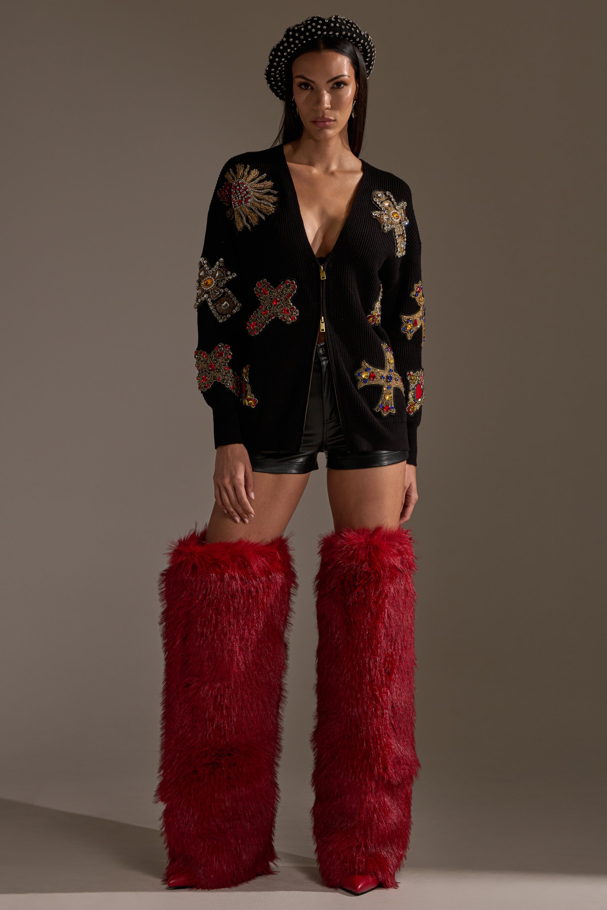 A woman wears the FEELING EXTRA EMBELLISHED SWEATER with colorful embroidered patches, black shorts, tall red furry boots, and a black beaded headband, standing against a plain background. 6