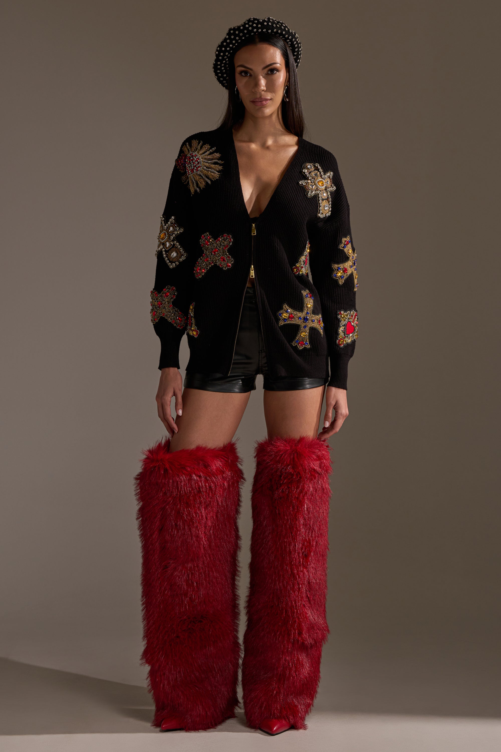 A woman faces forward in the FEELING EXTRA EMBELLISHED SWEATER featuring colorful crosses, paired with black leather shorts, a studded black beret, and tall bright red furry boots against a plain neutral background. 7