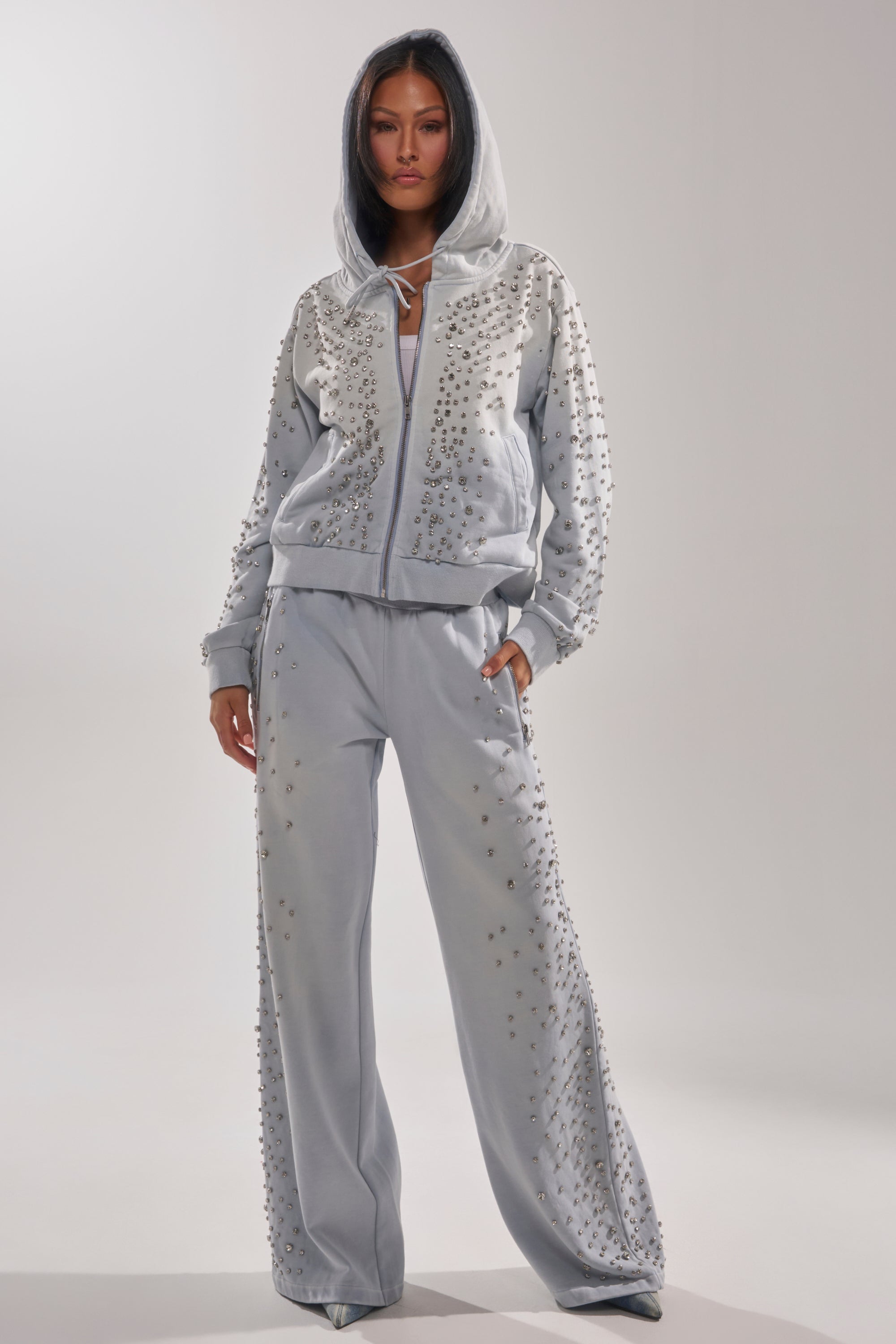 A woman wears the OH HOT DAMN EMBELLISHED JOGGER, a light gray rhinestone tracksuit with silver studs, including a zip-up jacket and wide-leg pants. She stands confidently with one hand in her pocket against a neutral backdrop. 0