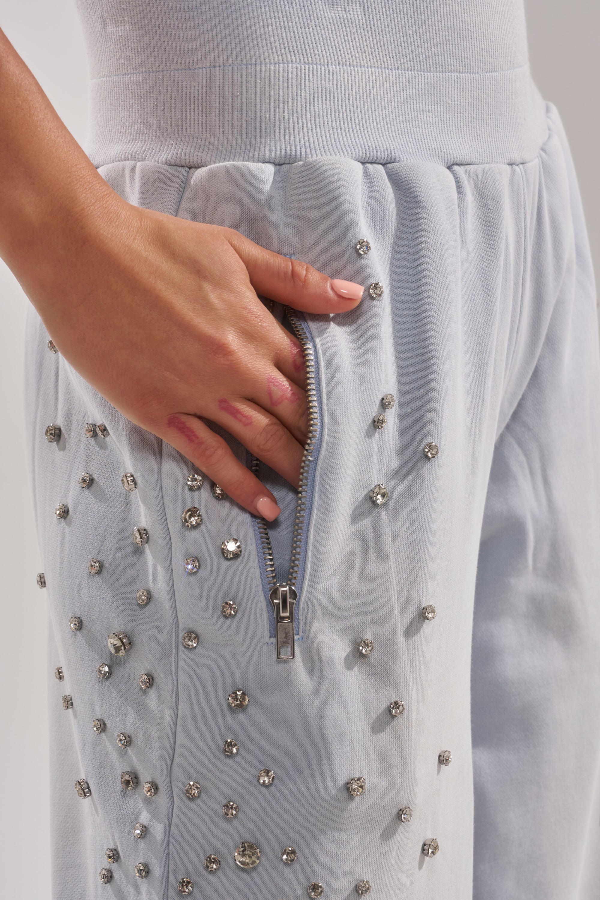 A hand partially inside the zippered pocket of the OH HOT DAMN EMBELLISHED JOGGER, light blue pants adorned with small, round rhinestones. 9