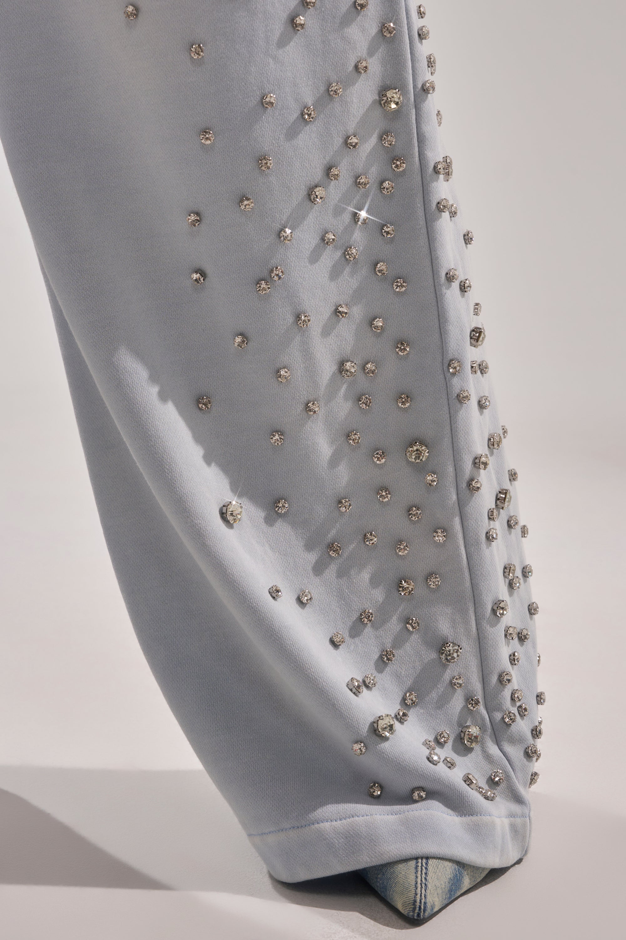 Close-up of the OH HOT DAMN EMBELLISHED JOGGER, showcasing diagonal sparkling rhinestones on light blue fabric, partially draped over a pointed-toe denim shoe against a neutral background. 10