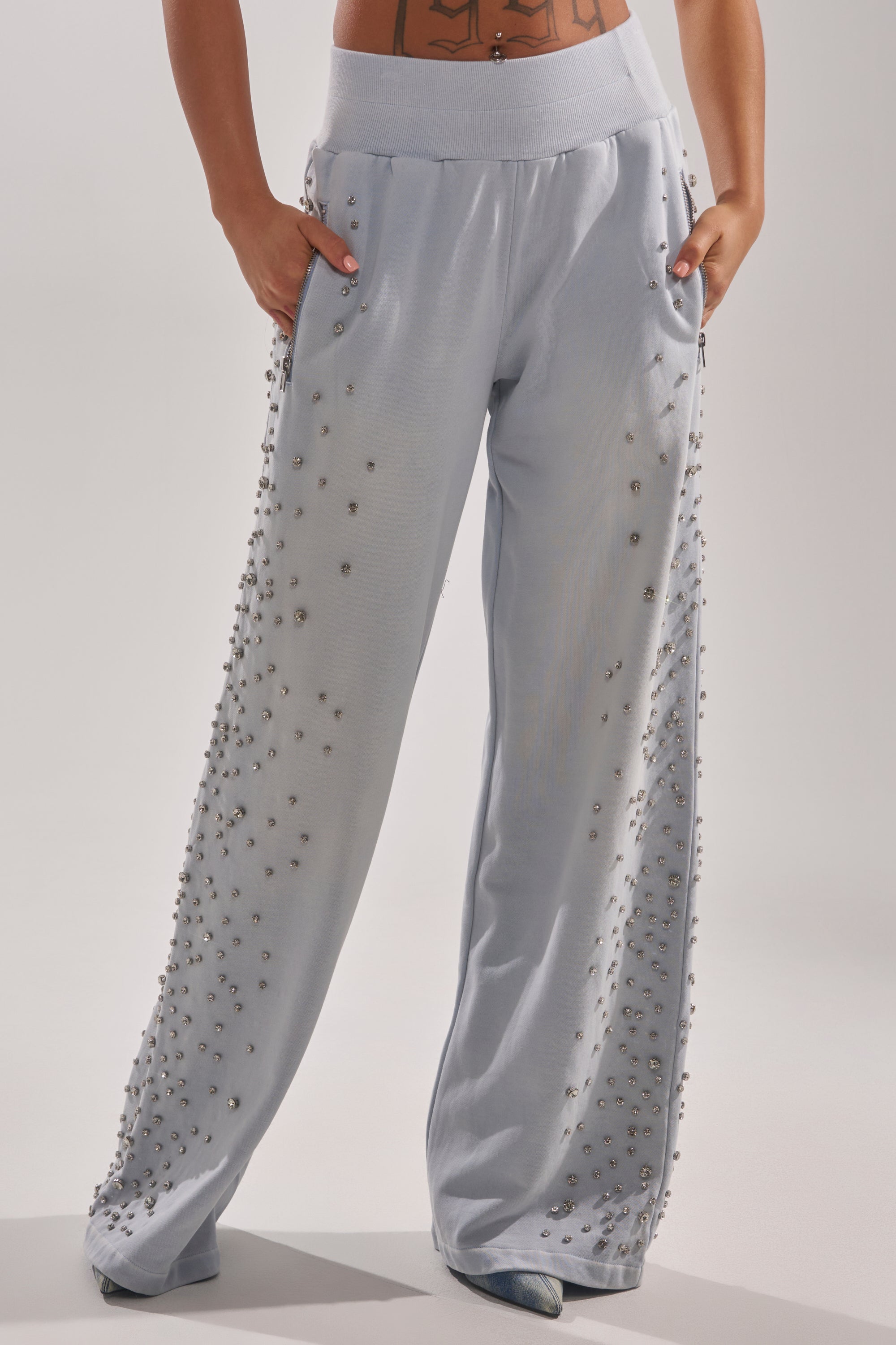 Someone stands against a plain background, hands in pockets, wearing the OH HOT DAMN EMBELLISHED JOGGER—light blue joggers decorated with small beads along the sides. 2