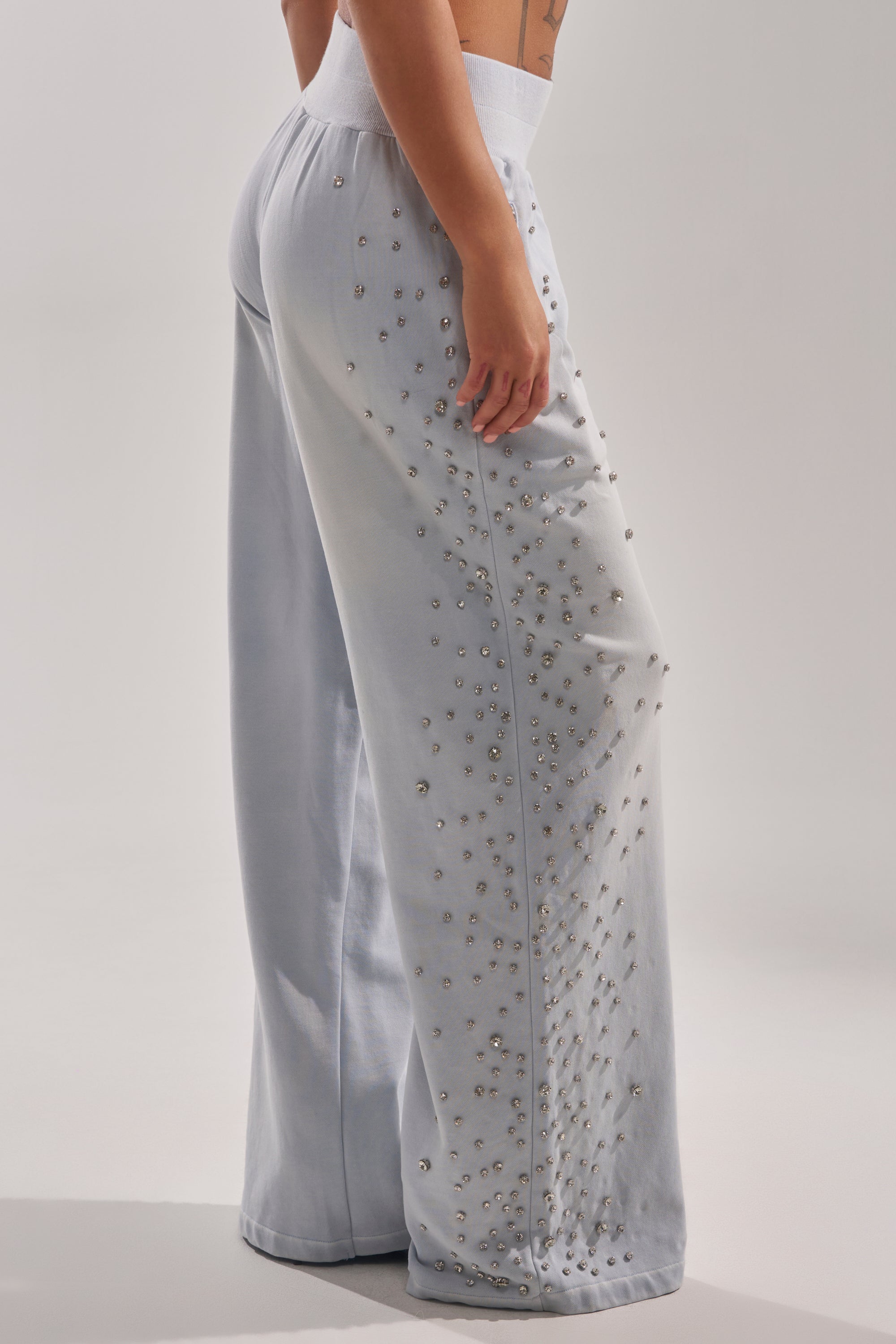 Someone stands against a plain light background wearing the OH HOT DAMN EMBELLISHED JOGGER—high-waisted, wide-leg sweatpants in light gray, featuring small silver studs along the sides. 3
