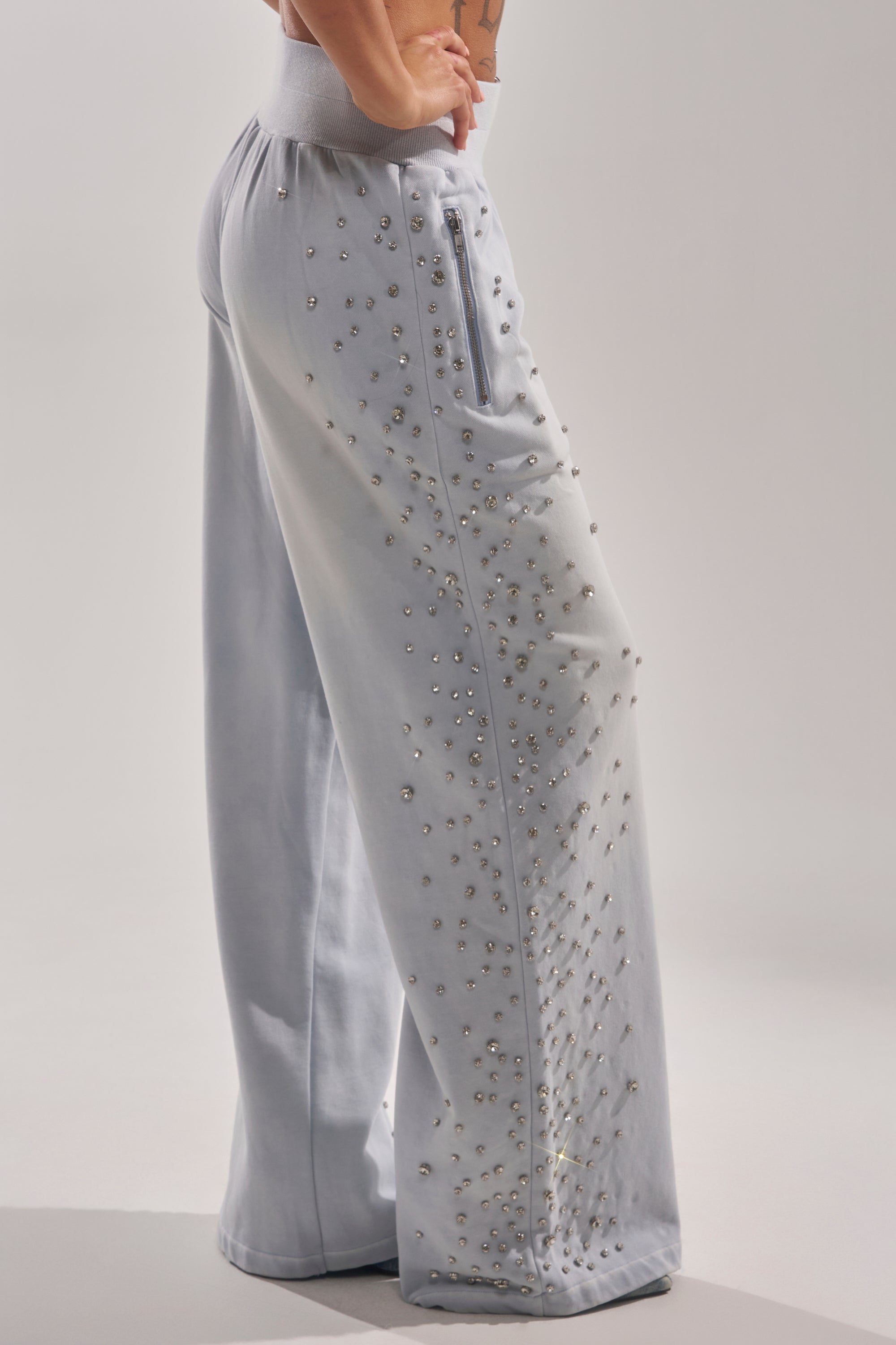 Someone models the OH HOT DAMN EMBELLISHED JOGGER—light gray, wide-leg sweatpants adorned with shiny studs on the front and side—against a plain, light background. 4