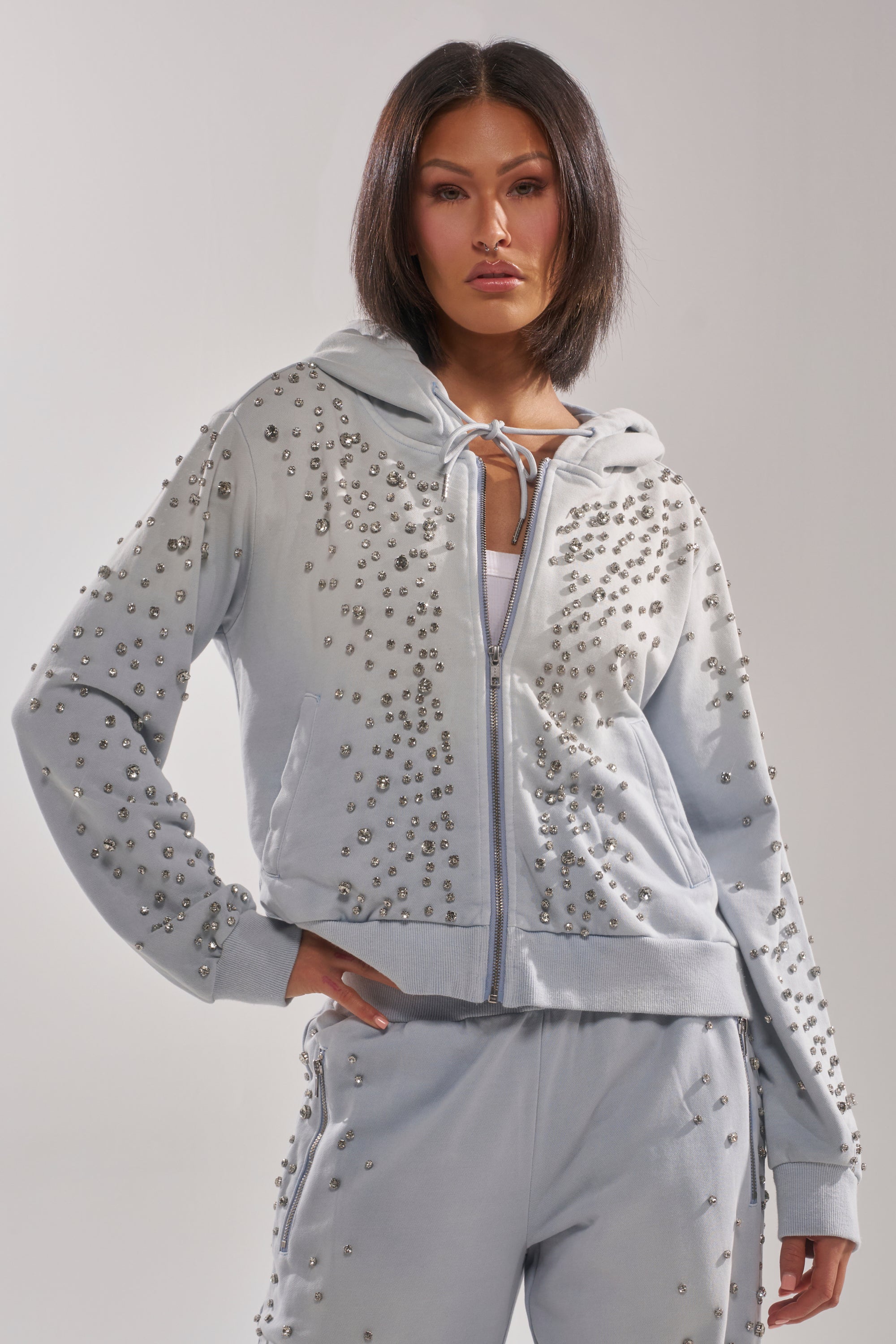 A woman with straight dark hair confidently poses in the OH HOT DAMN EMBELLISHED ZIP UP SWEATSHIRT and matching sweatpants, both adorned with clusters of silver studs, against a plain light background. 0