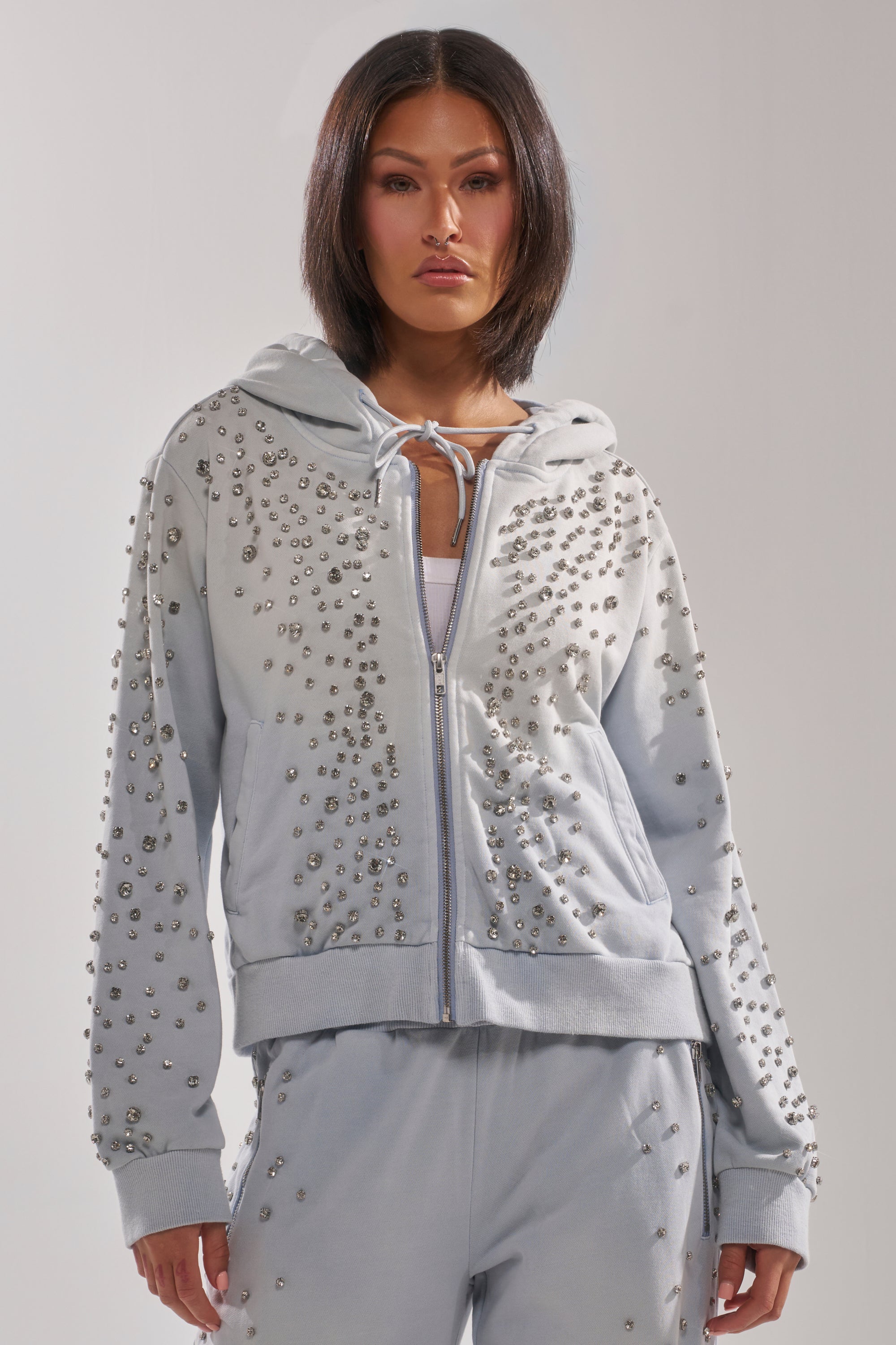 A woman wears the OH HOT DAMN EMBELLISHED ZIP UP SWEATSHIRT by AKIRA Label and matching pants, each adorned with clusters of metallic studs, as she stands facing forward against a plain background with a neutral expression. 1