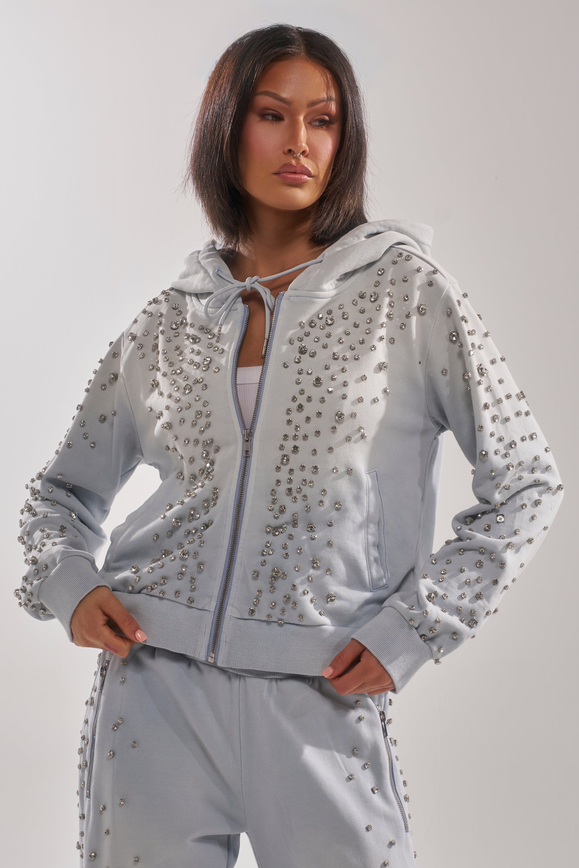 A woman wears the OH HOT DAMN EMBELLISHED ZIP UP SWEATSHIRT in light gray with matching studded pants, standing with her hands in her pockets and looking to the side with a neutral expression. 2