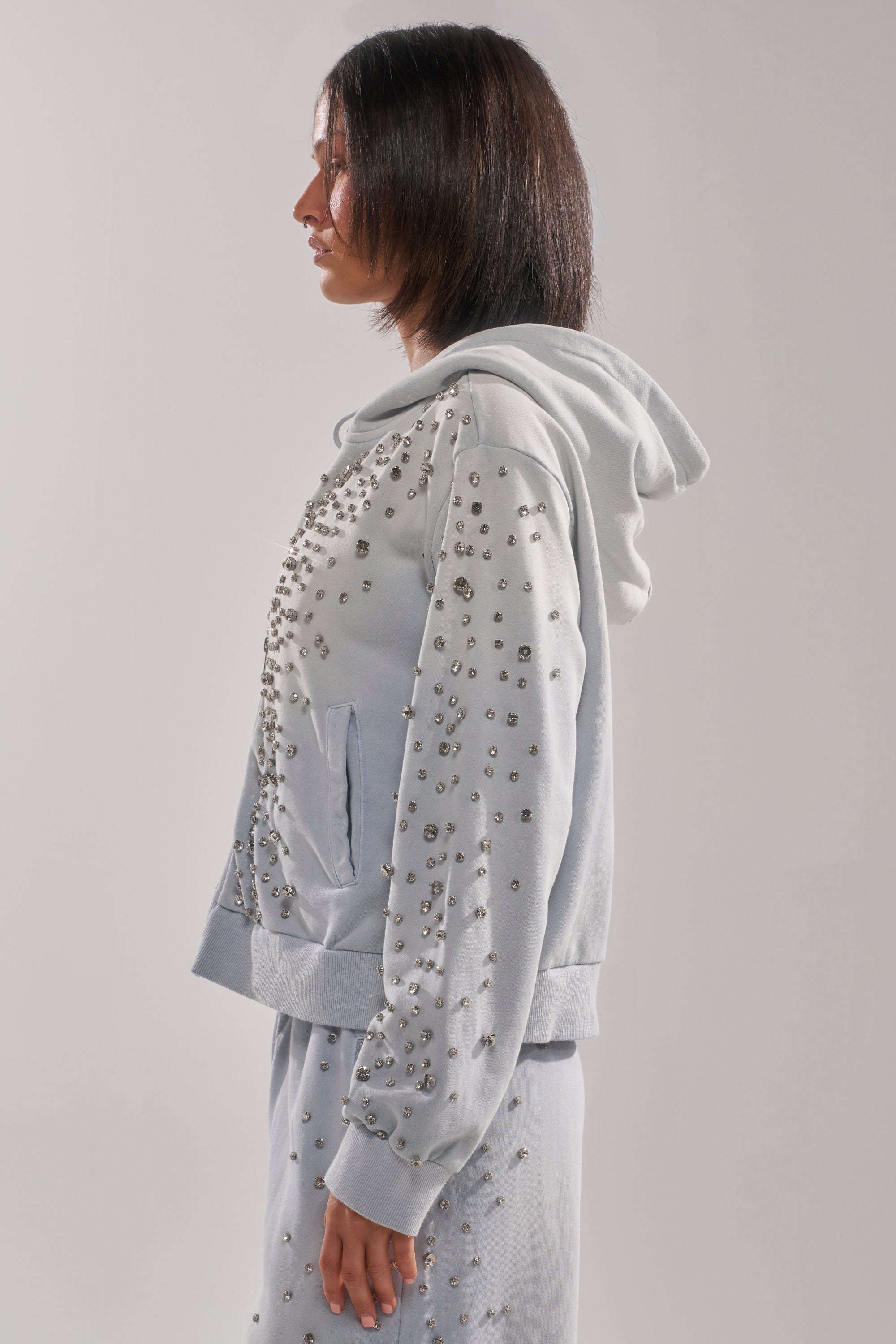 A person with straight, shoulder-length dark hair is shown in profile wearing the OH HOT DAMN EMBELLISHED ZIP UP SWEATSHIRT, detailed with small metallic studs, against a plain light background. 3