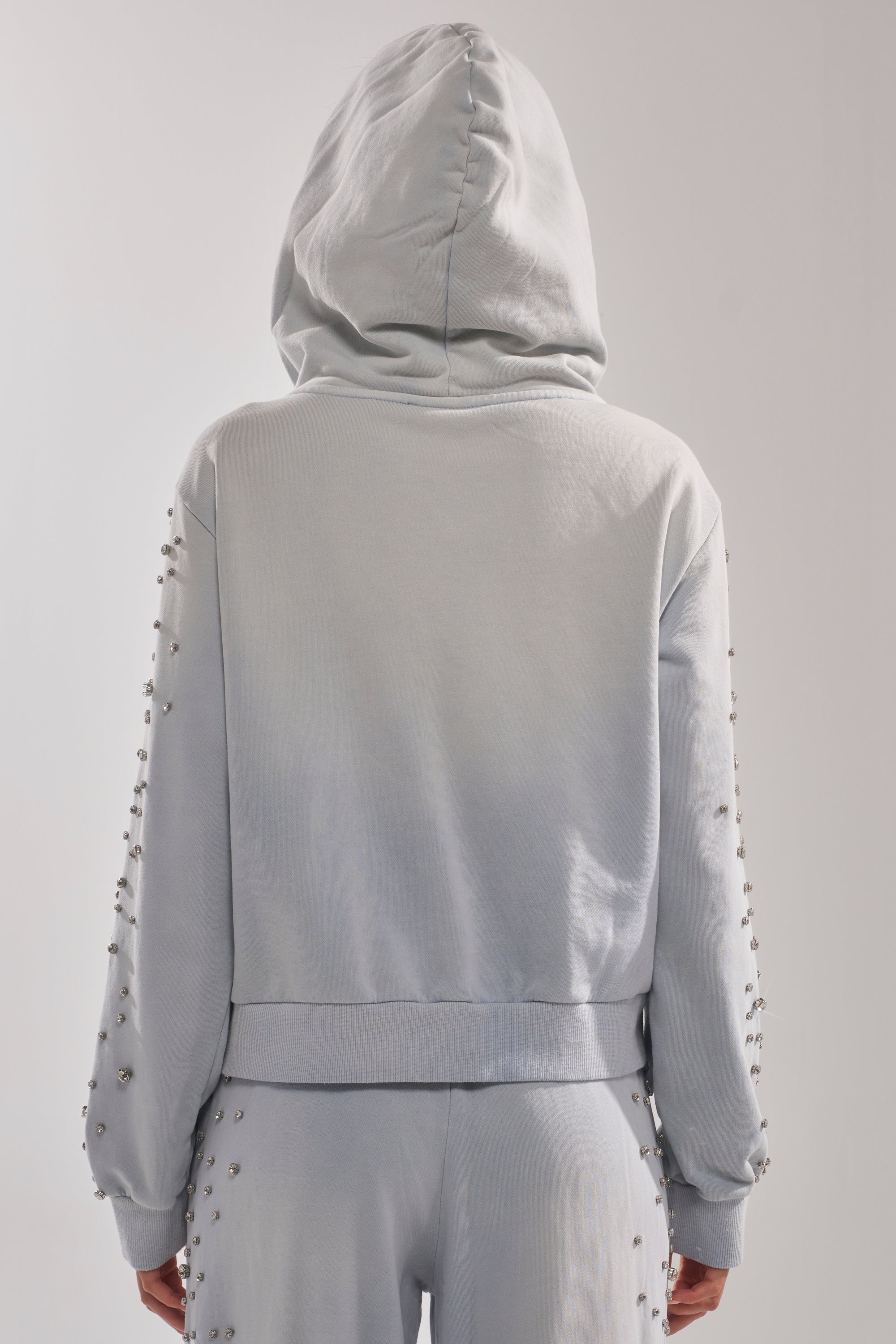 A person, seen from the back, wears the AKIRA Label OH HOT DAMN EMBELLISHED ZIP UP SWEATSHIRT in light gray with the hood up; both the sweatshirt and sleeves feature small round stud decorations. 4