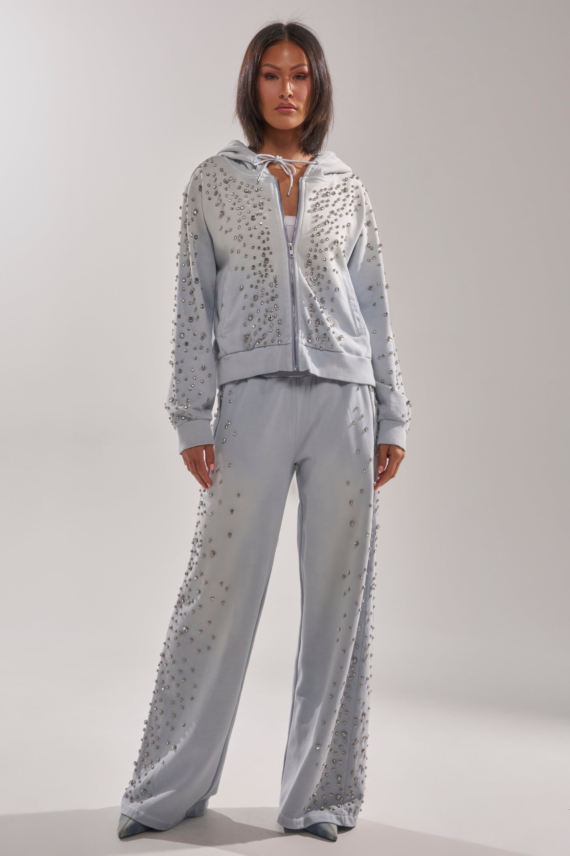 A woman with straight dark hair and a neutral expression faces forward, wearing the AKIRA Label OH HOT DAMN EMBELLISHED ZIP UP SWEATSHIRT and matching studded wide-leg pants against a plain light background. 6