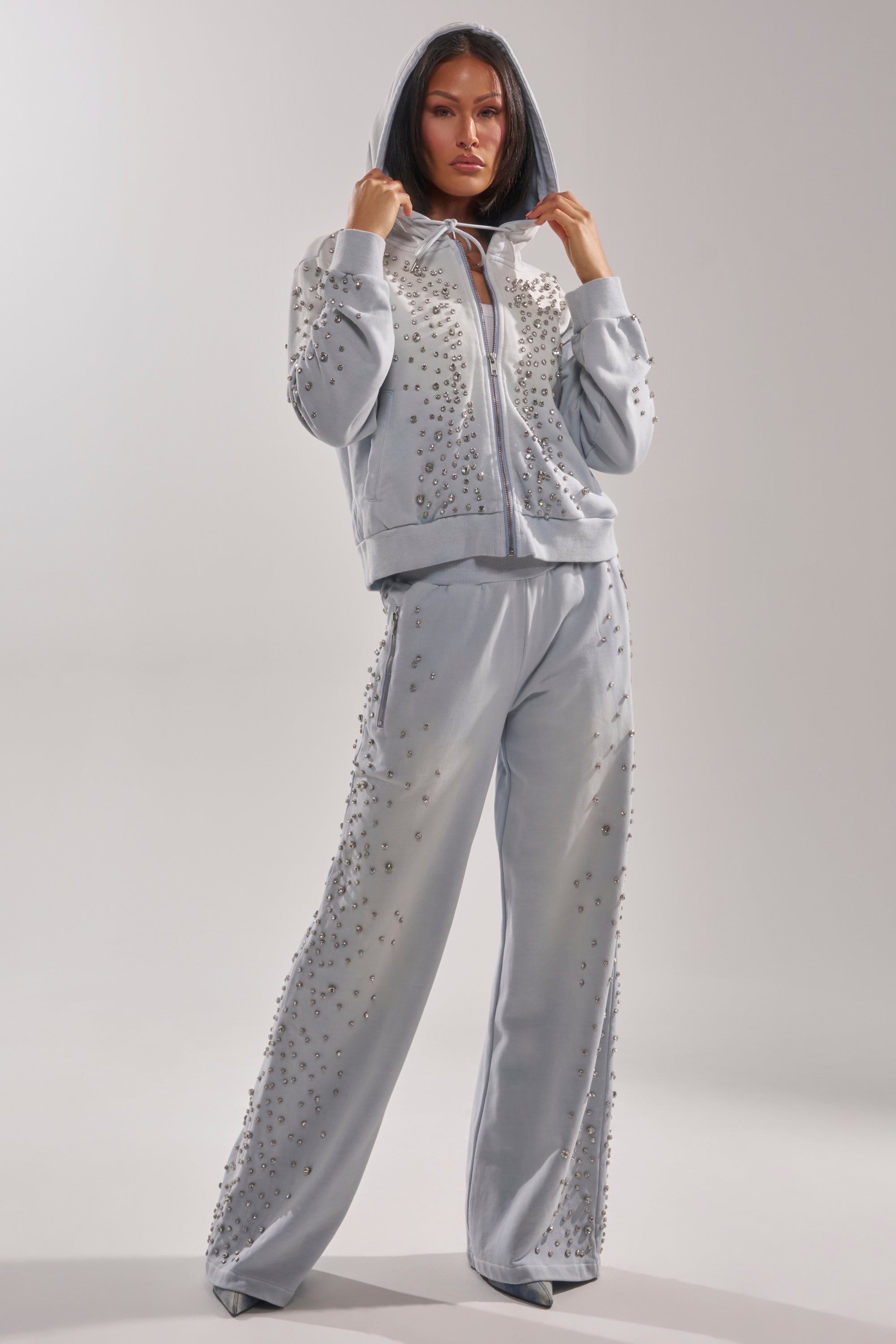 A woman poses confidently in the light gray OH HOT DAMN EMBELLISHED ZIP UP SWEATSHIRT, featuring sparkling beads and rhinestones, hood up and hands holding the edges against a plain background. 8