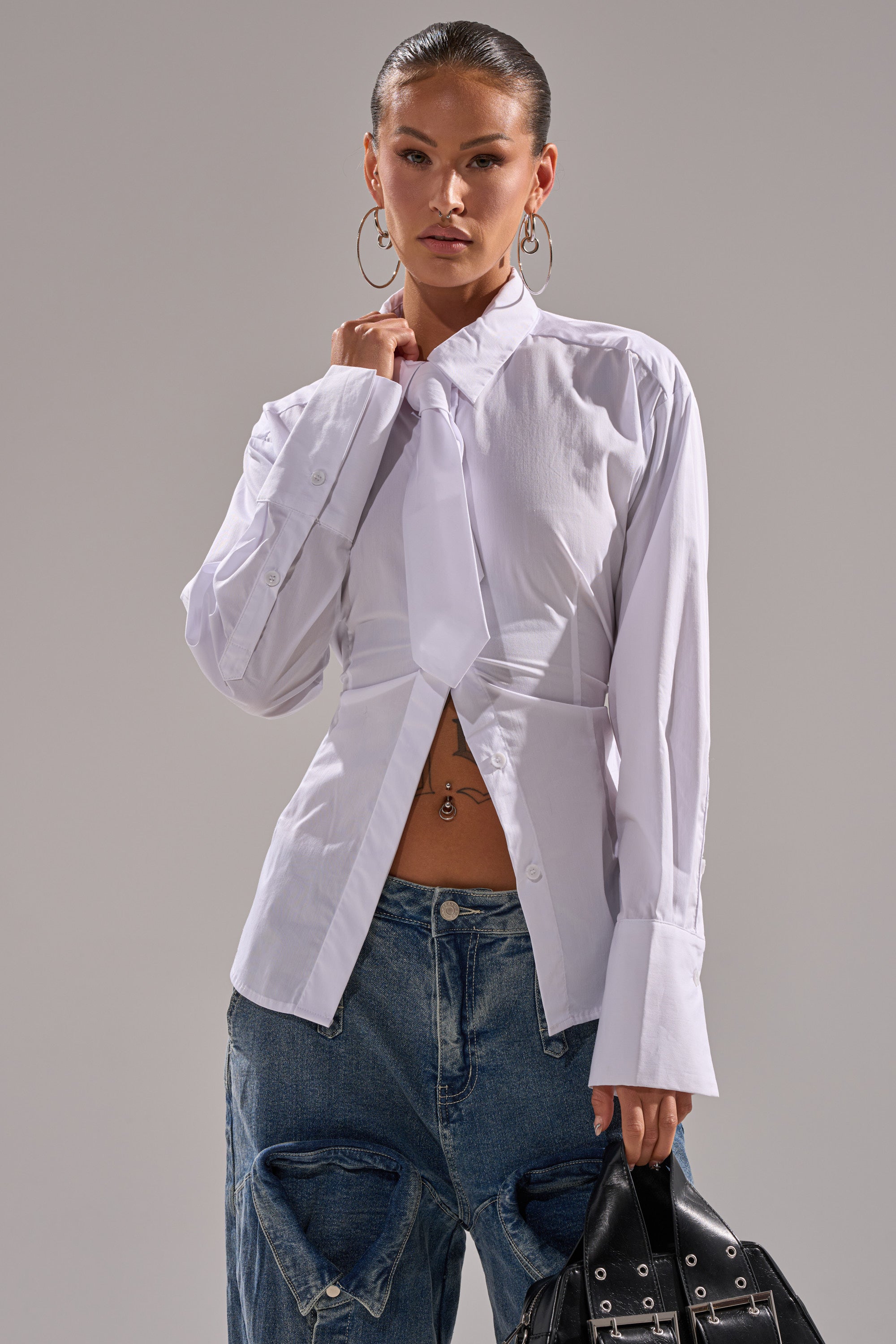 A woman styles the BUSINESS BUTTON DOWN with oversized cuffs and an open collar, pairing it with distressed blue jeans, a black handbag, large hoop earrings, and a visible belly button piercing.