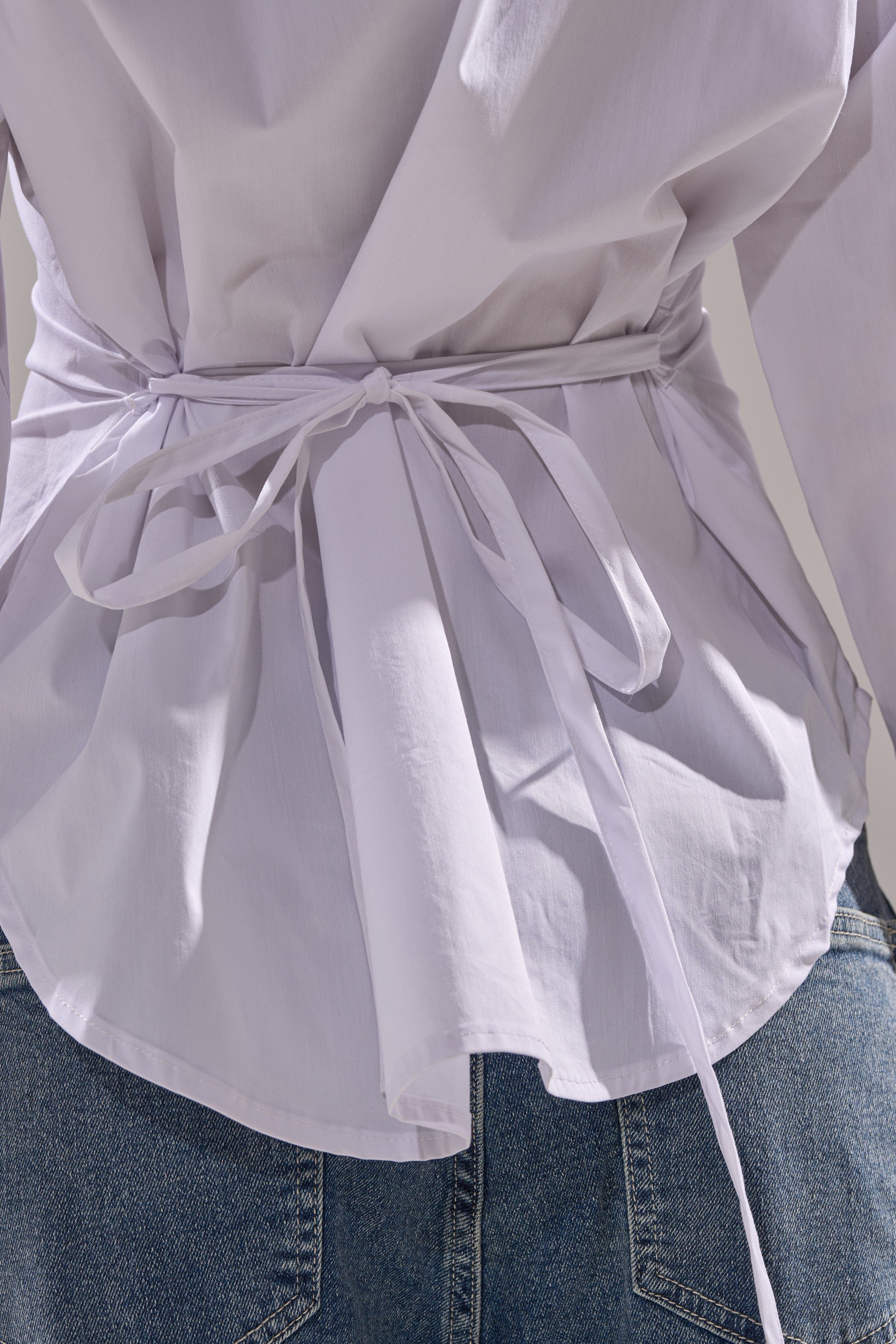 A close-up of a person wearing the BUSINESS BUTTON DOWN, a light cotton blouse tied at the waist with a thin belt and paired with blue denim jeans. The photo, taken from the back, highlights the blouse’s fabric and bow detail.
