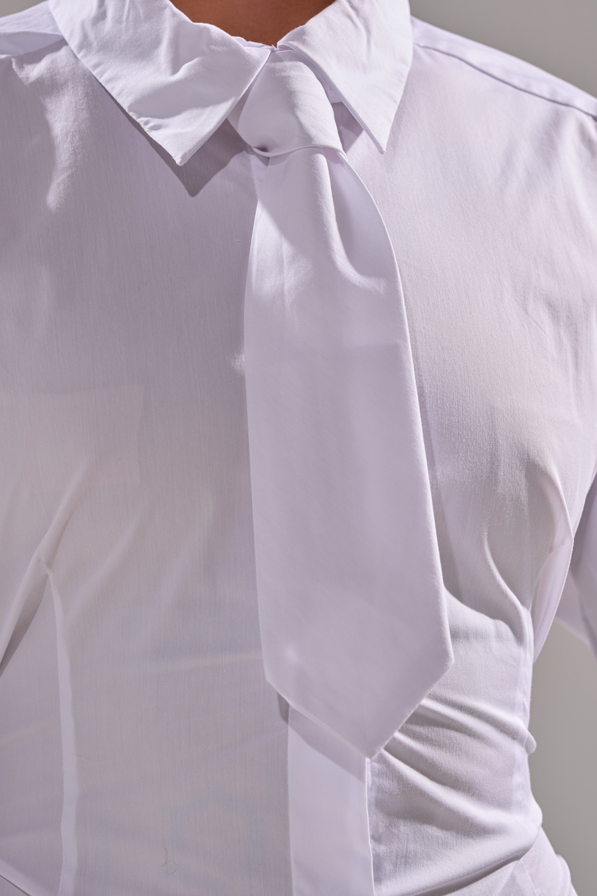 A person wears the BUSINESS BUTTON DOWN, a sheer white blouse with a white tie; its slightly translucent cotton fabric subtly reveals skin tone beneath, focusing on torso and clothing details.