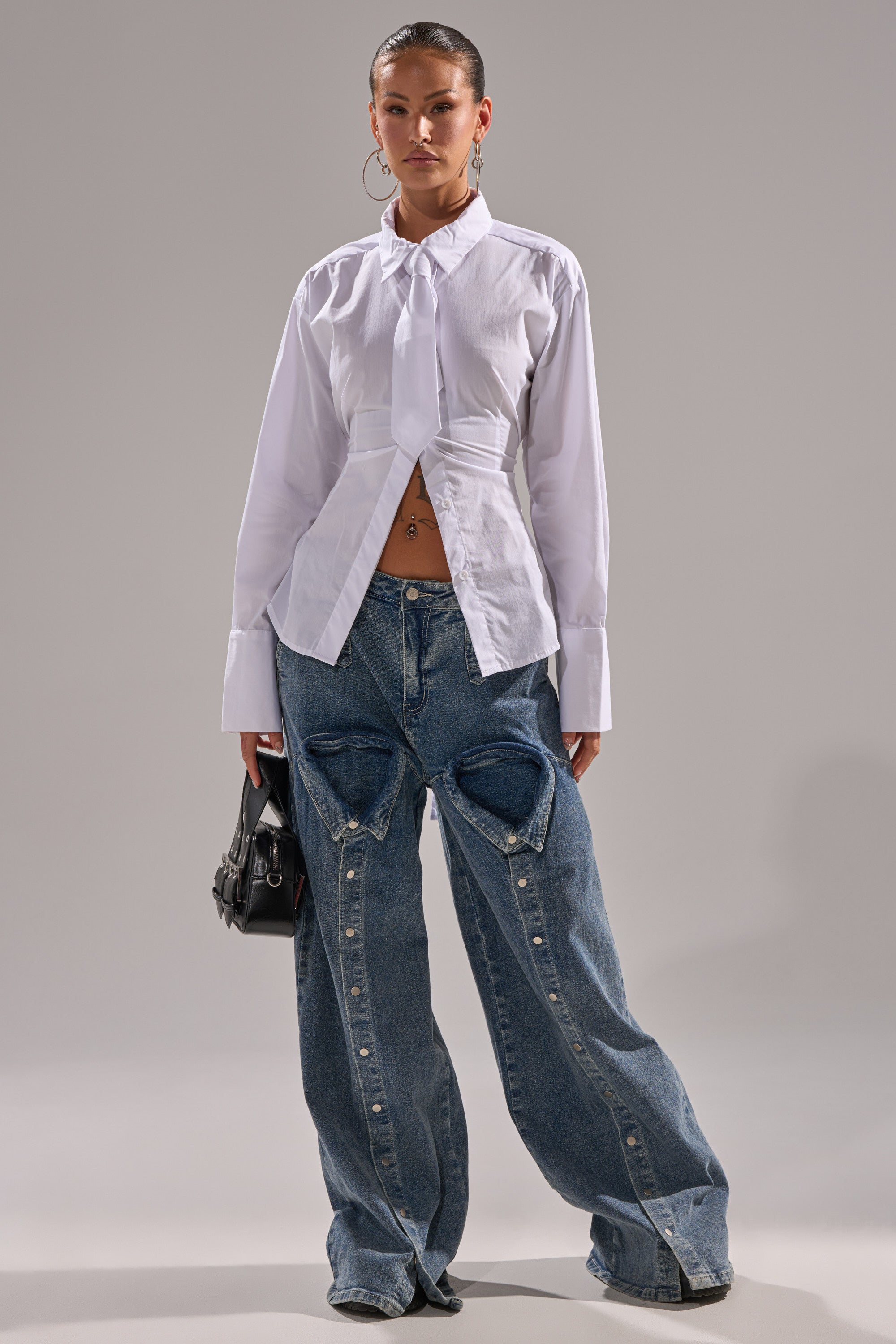 Wearing the BUSINESS BUTTON DOWN—a white cotton blouse with an asymmetrical hem and oversized cuffs—paired with baggy blue jeans with heart designs, a black handbag, and slicked-back hair.